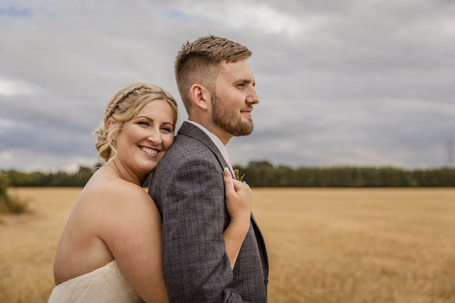 A bride and groom embrace in the countryside at Shustoke Barn. The bride wears a white strapless gown and the groom a grey checked suit.