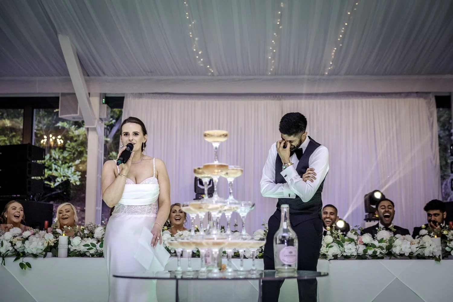 A bride is visibly roasting her new husband as the bridal party looks on in shock and laughter. The bride wears a boutique gown and the groom wears black tie. They stand behind a four tied champagne fountain containing pink fizz.