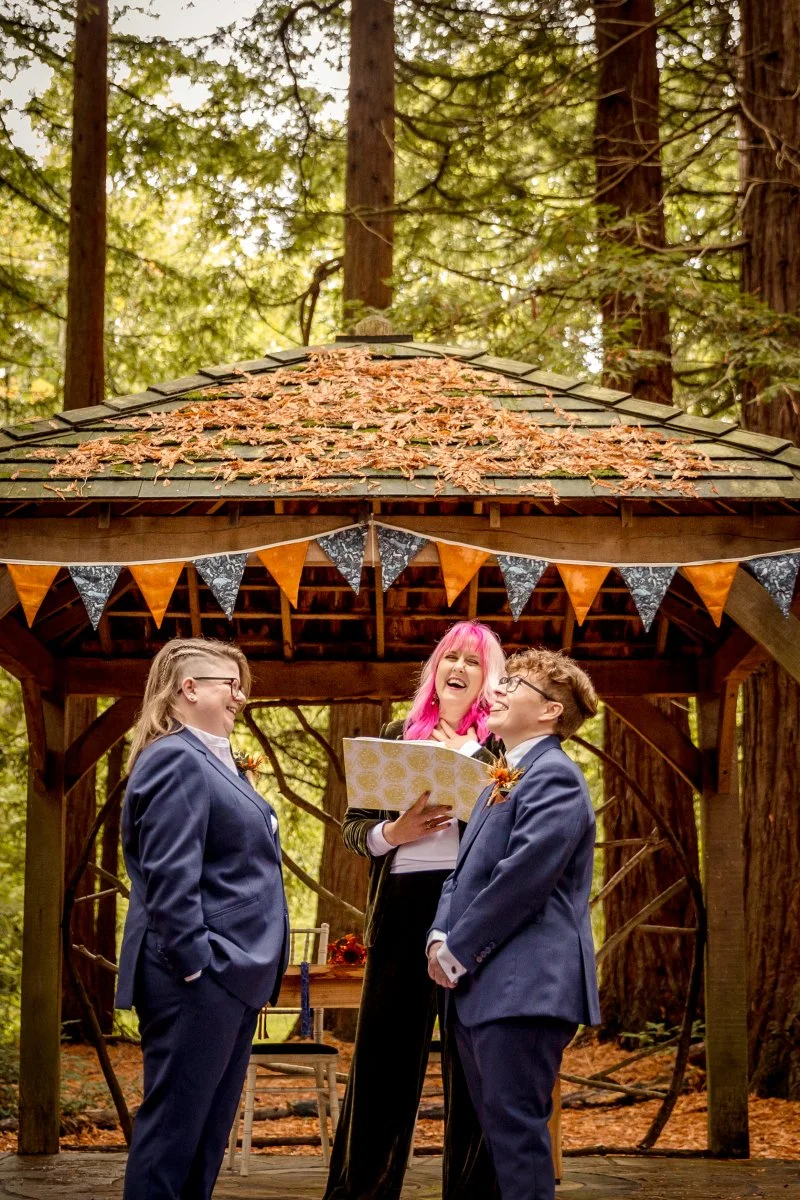 An LGBT laugh at the woodland alter at Two Woods Estate, Pulborough, with their pink haired wedding Celebrant. They are surrounded by burnt orange and navy bunting, framed well with the golden leaves.
