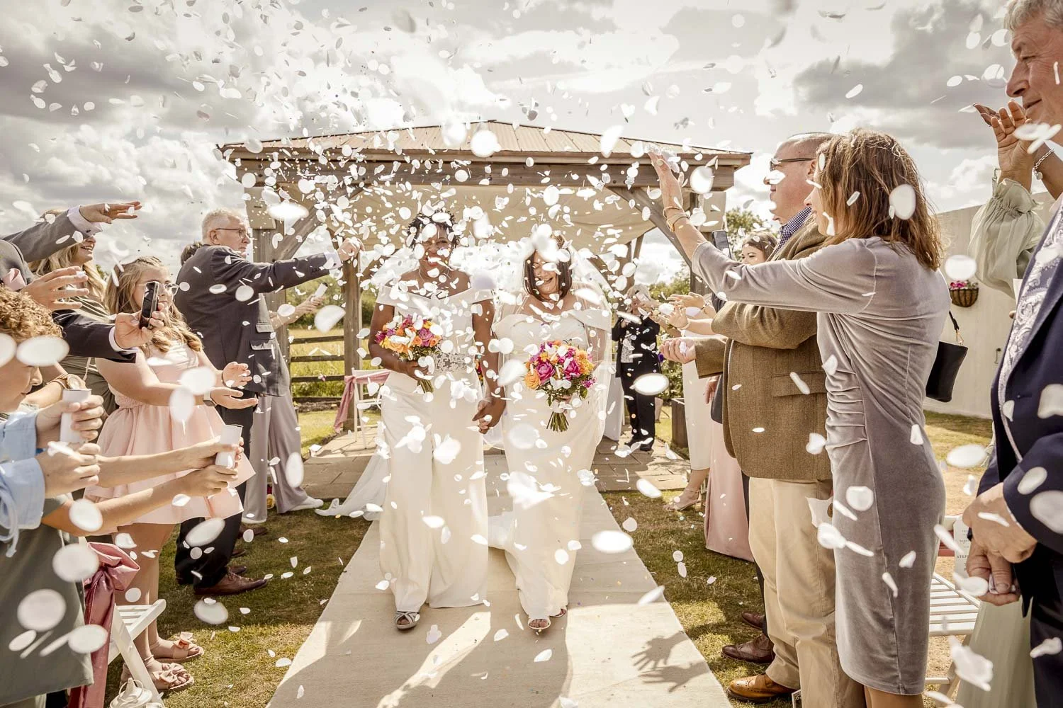 White confetti surrounds brides at their outdoor wedding ceremony at Badgers Mount Hotel in Elmsthorpe Leicestershire