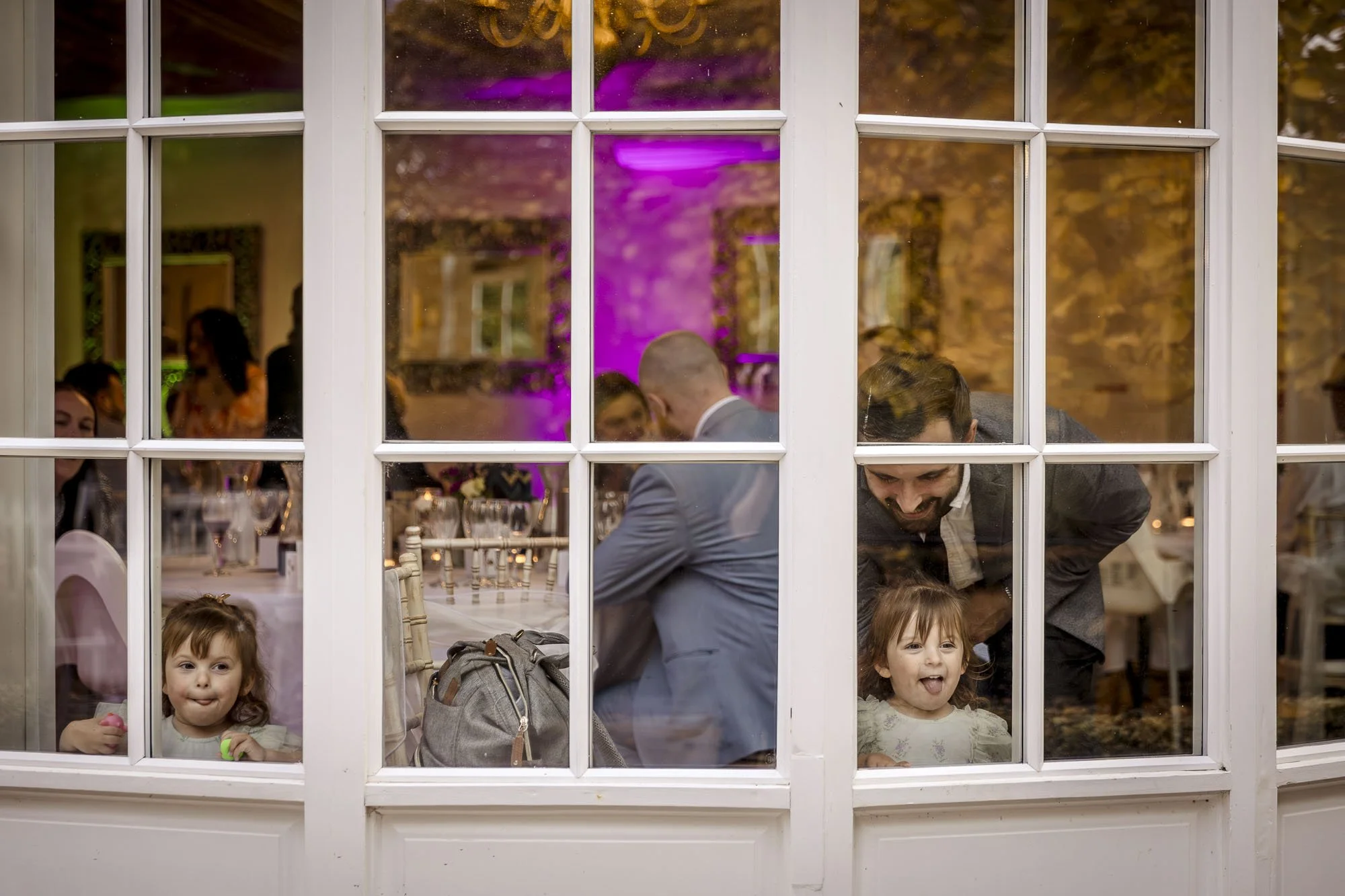 Two girls watch from the window sticking their tongues out as they watch the bride and groom have their portraits taken