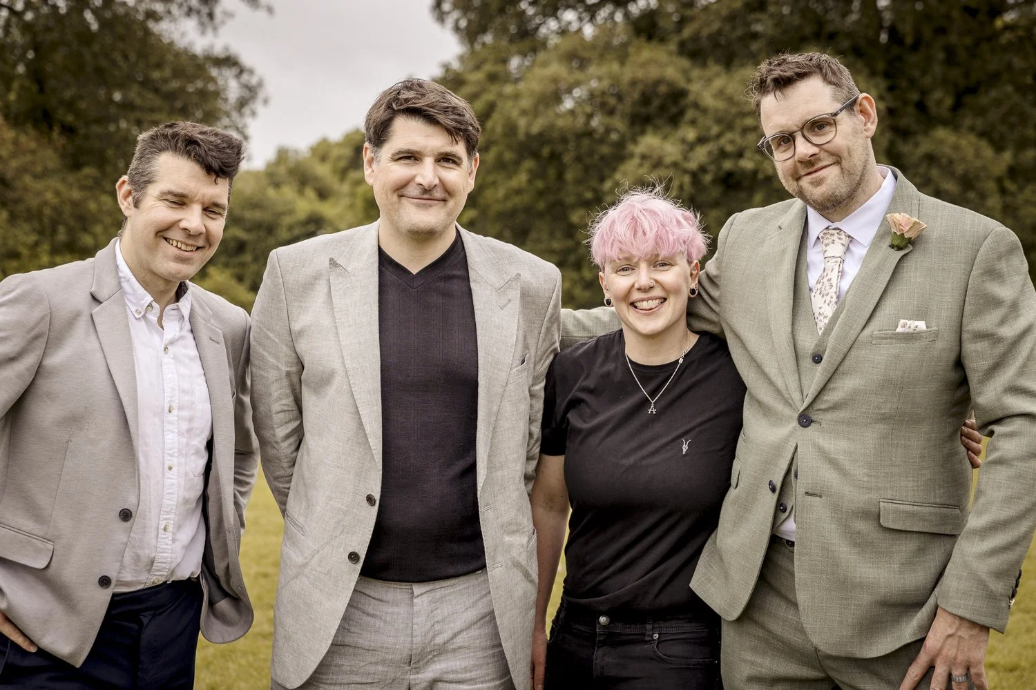 Amie Seal a pink haired wedding photographer stands with three brothers on the finale of their weddings. They wear neutral coloured suits and are surrounded by trees. Amie meanwhile, wears her signature black attire