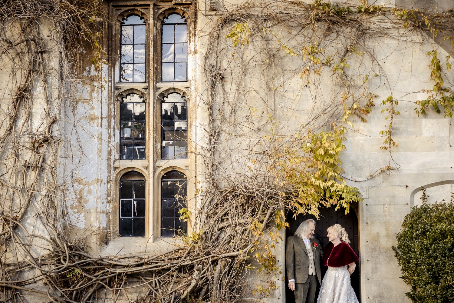 A mature bride and groom look at each other smiling with cheeky faces within the doorway of Thornburn Castle near Bristol. The impressive background shows ornate 16th century windows and vines crawling the stone castle walls