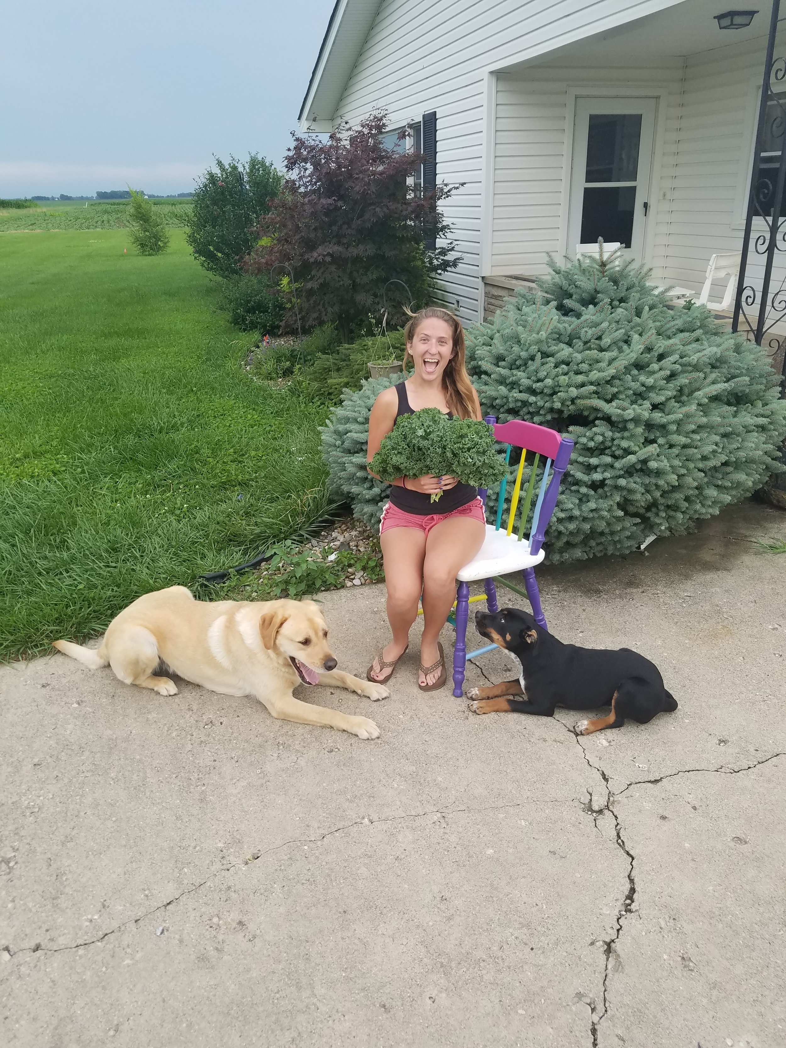 Jessica posing with her homegrown kale, pups, and the piece she is restoring.