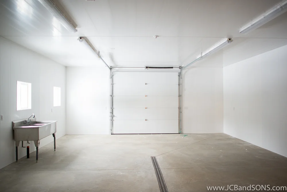 Interior of honey extraction room in Southgate with concrete floor, center drain, white vinyl wall and ceiling panels, and new overhead door.