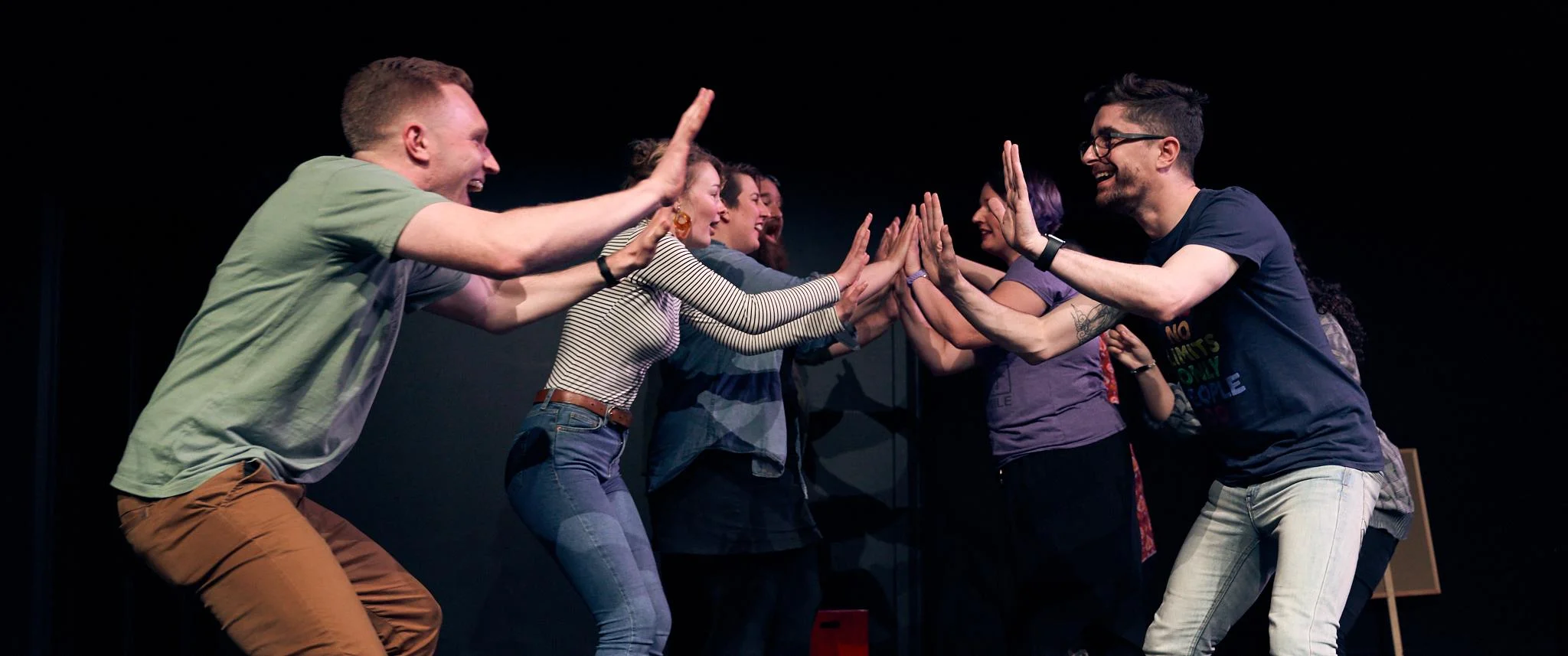 Image from The Protest, NZIF 2018. Eight improvisors are face each other in two lines, hands outstretched to high-ten each other. They are smiling.