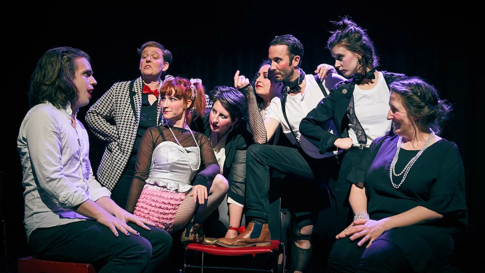 The Kit Kat Prov, photo by Joel Luscombe  Image description: One improvisor sitting facing a group of performers as if telling a story. The other improvisors are dressed in a shabby-chic caberet style, lots of red and black and white, and they hold a very Cabaret pose - finger snaps, legs up, leaning on each other.