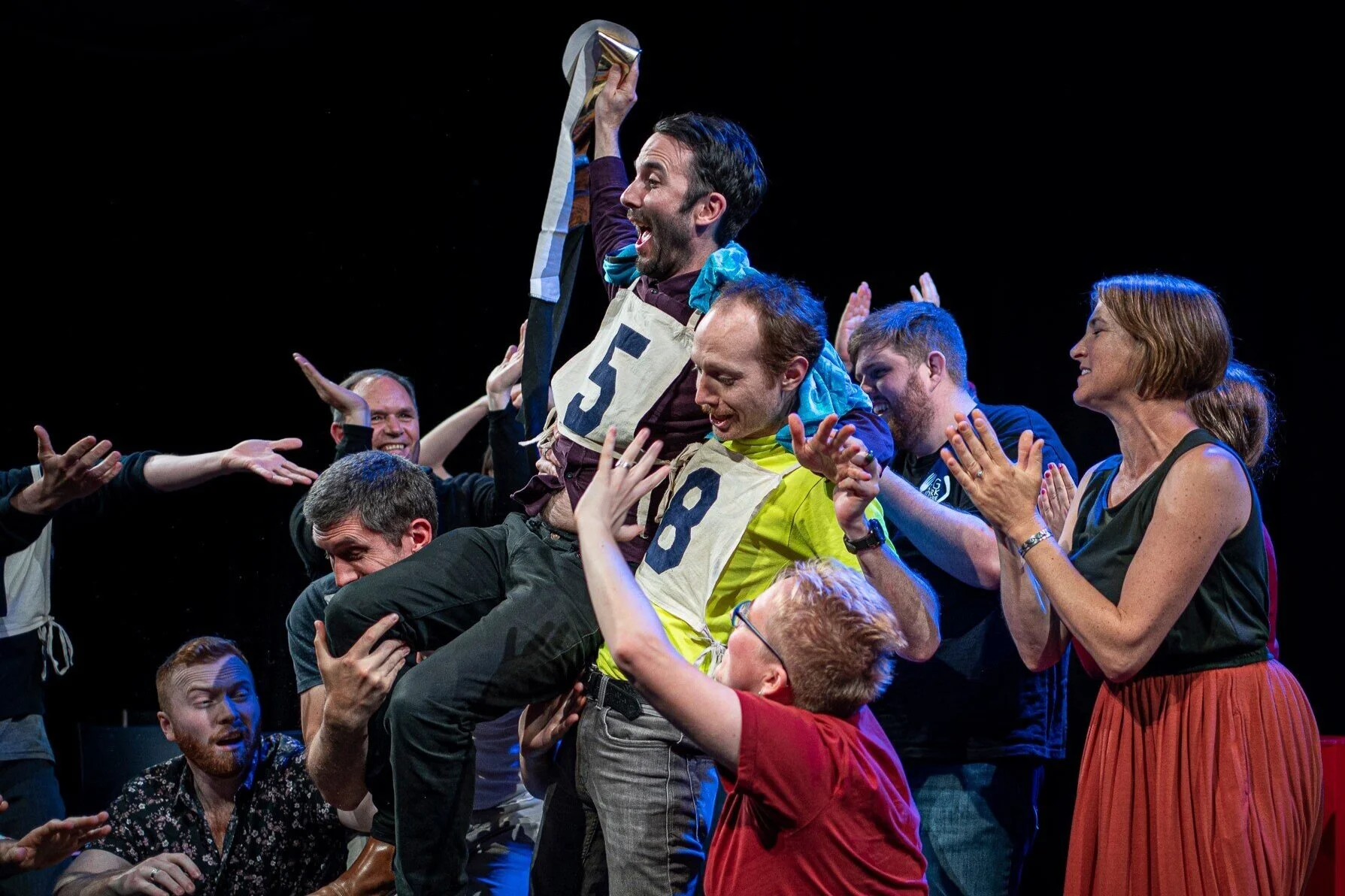 Image description: a group of improvisors wearing numbered bibs lift the winner of one of the heats of All-In All-Out Improv Bout on their shoulders, clapping and smiling