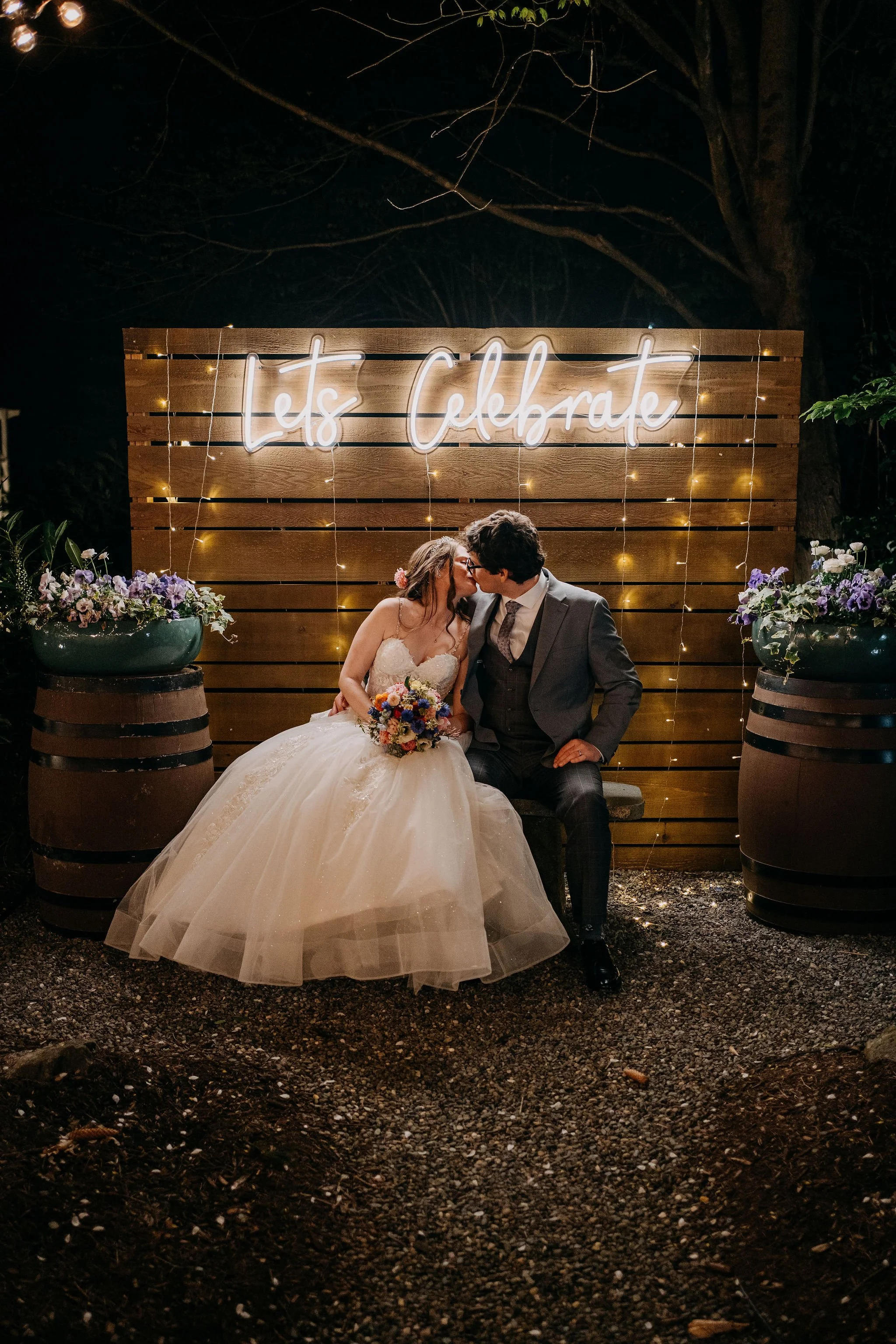 Bride and groom kissing in front of a rustic wooden backdrop and illuminated neon sign during their Grain House at the Olde Mill Inn wedding.