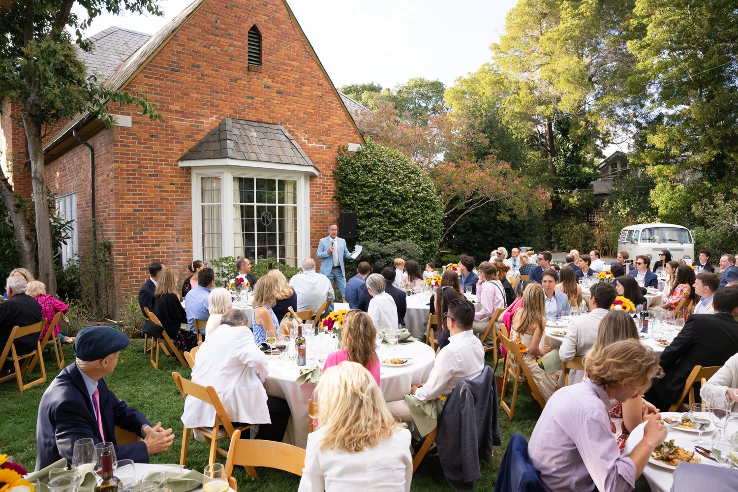 Outdoor gathering of people seated at round tables with white tablecloths, colorful floral centerpieces, food, and drinks, listening to a man in a light blue suit giving a speech in front of a brick house with large windows, surrounded by green trees