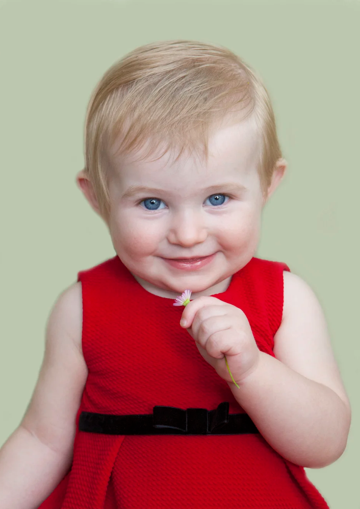 A young girl with blue eyes and blonde hair wearing a red dress with a black bow, holding a small pink flower, smiling at the camera against a light green background.