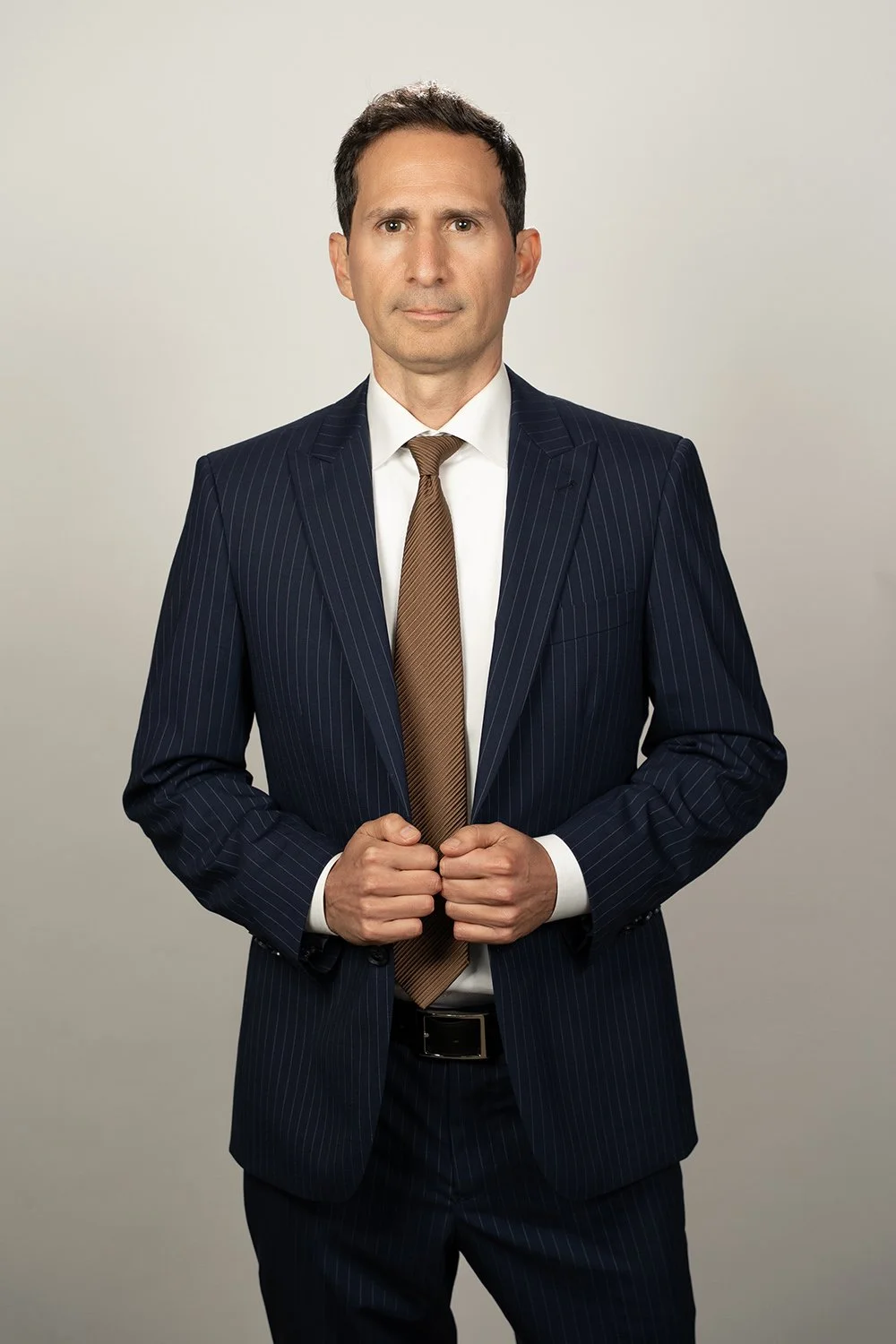 A man in a navy pinstripe suit with a white shirt and brown tie, holding the front of his suit jacket, standing against a plain light-colored background.
