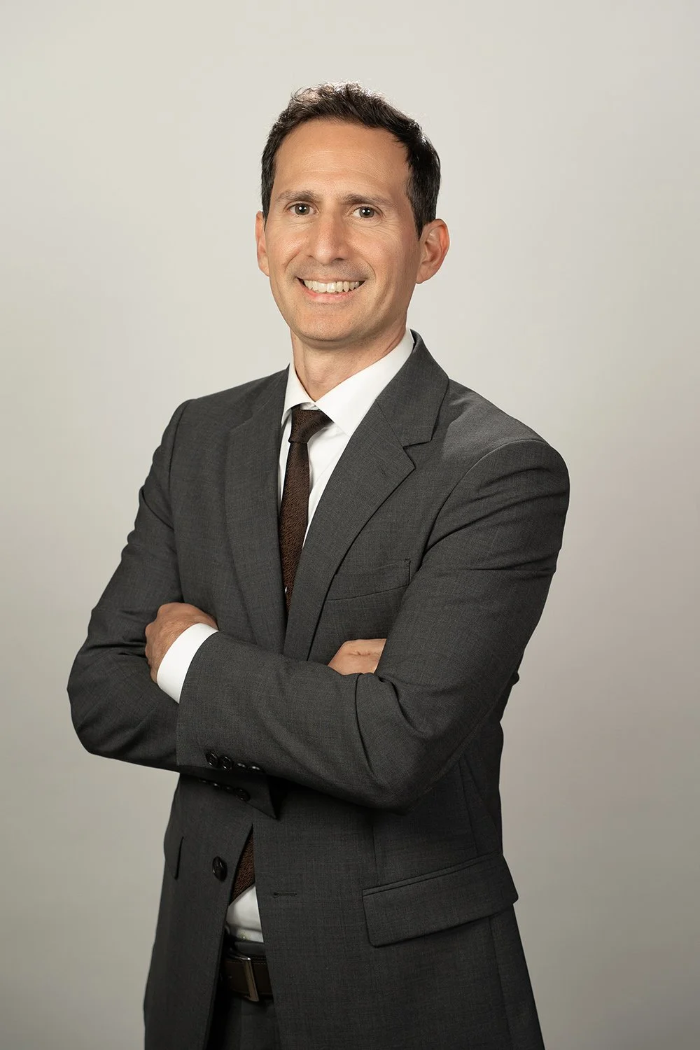 A man in a gray suit with a white shirt and dark tie standing with arms crossed smiling in front of a plain light background.