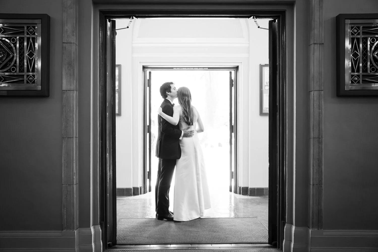 A black and white photo of a bride and groom sharing a kiss at the entrance of a building, possibly during a wedding or wedding photoshoot.