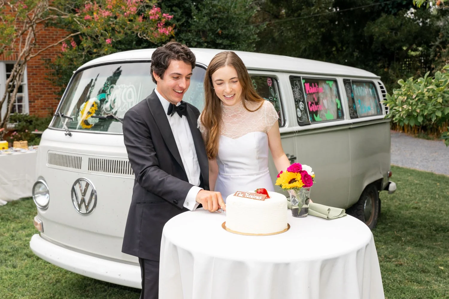 A bride and groom in wedding attire cutting a wedding cake outdoors, with a vintage Volkswagen van decorated with colorful signs in the background.