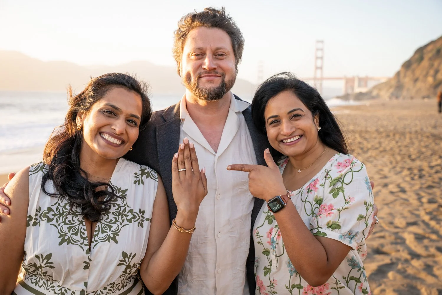 Three people on a beach during sunset, smiling and showing support for same-sex marriage with a woman displaying her ring and another woman pointing to it.