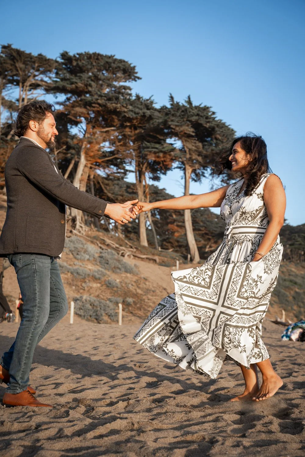 A man and woman are on the beach, dancing and holding hands. The man wears a dark blazer and jeans; the woman wears a sleeveless patterned dress. They are smiling, with trees and a clear sky in the background.