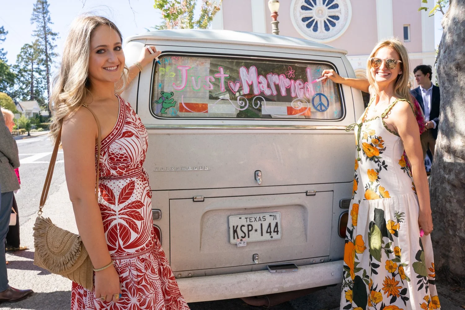 Two women standing near a white Volkswagen van decorated with a colorful sign reading 'Just Married' and peace symbols, at a wedding celebration outdoors.