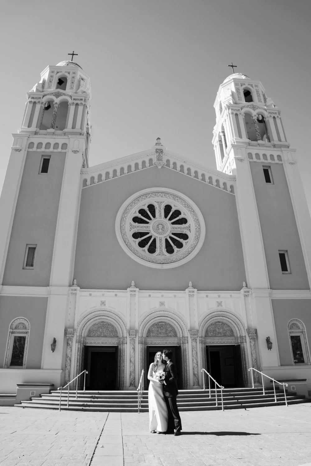 A couple in wedding attire standing in front of a large church with twin bell towers, a rose window, and ornate entrance doors.