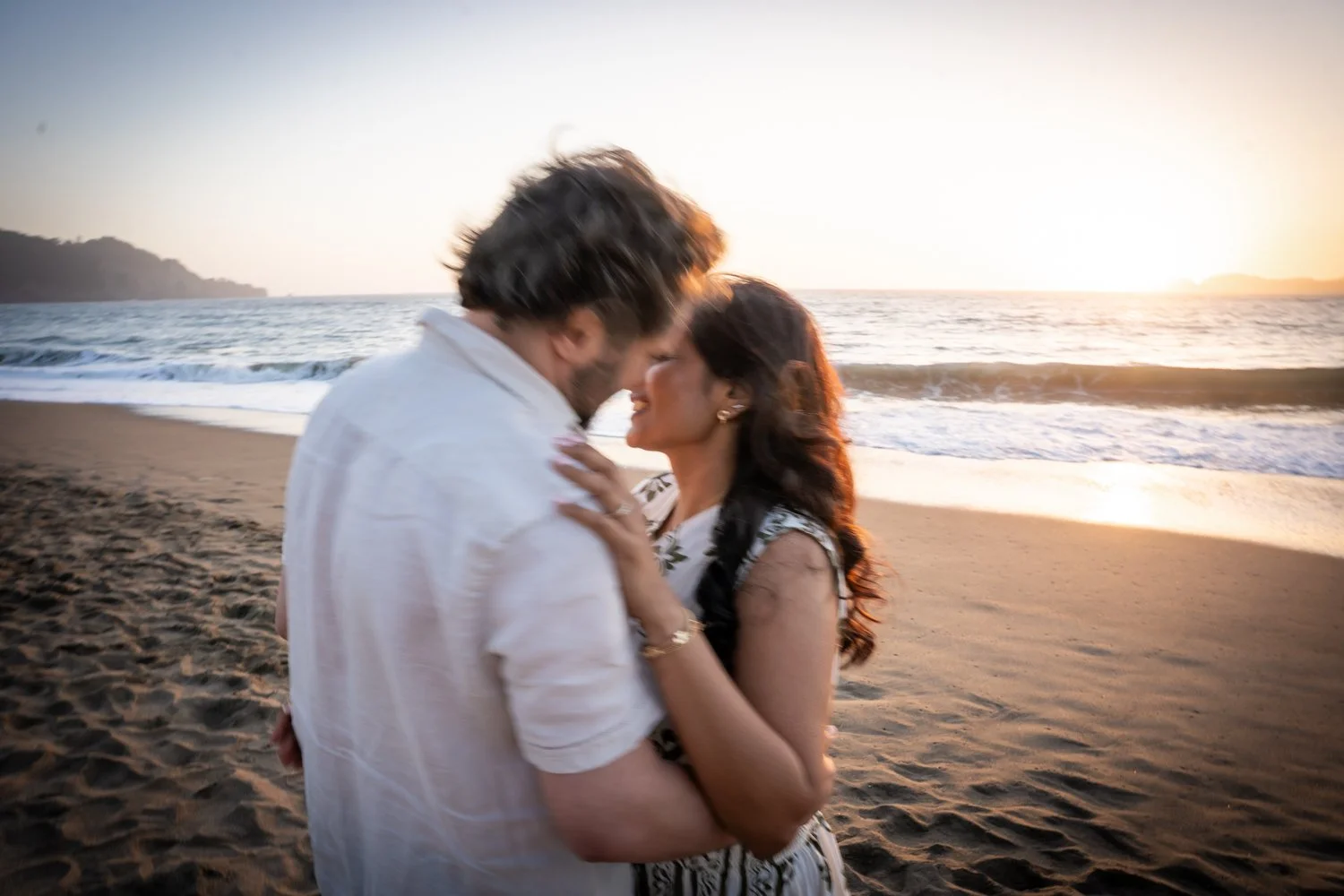 A couple embraces closely on a beach at sunset, with the ocean and a distant shoreline in the background.