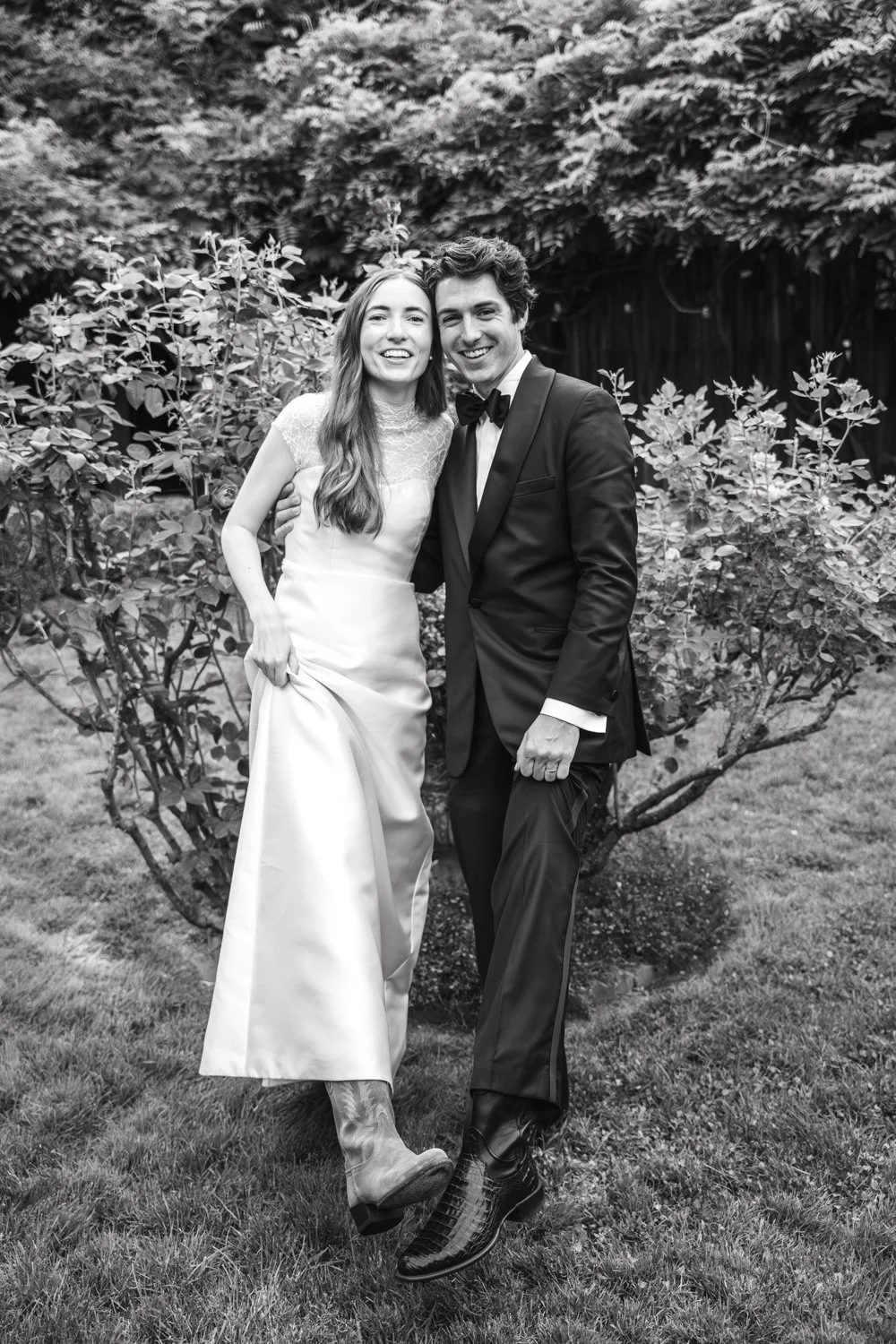A smiling couple in formal attire, the woman in a white gown with lace details and cowboy boots, the man in a tuxedo with a bow tie, standing on grass in front of bushes and trees.