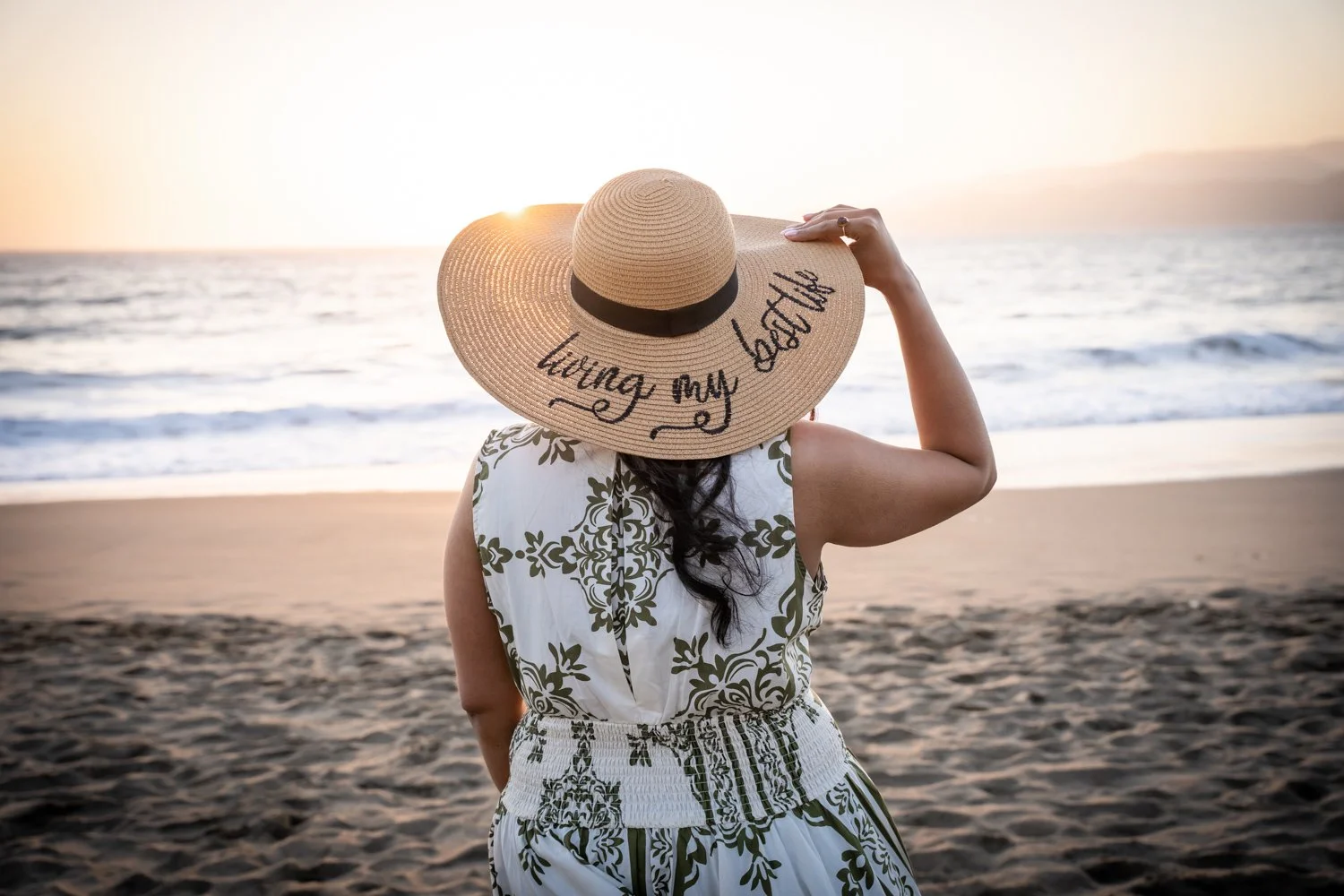 A woman on a beach during sunset, wearing a wide-brimmed straw hat with the words "living my best life" written on it and a white dress with a green floral pattern.