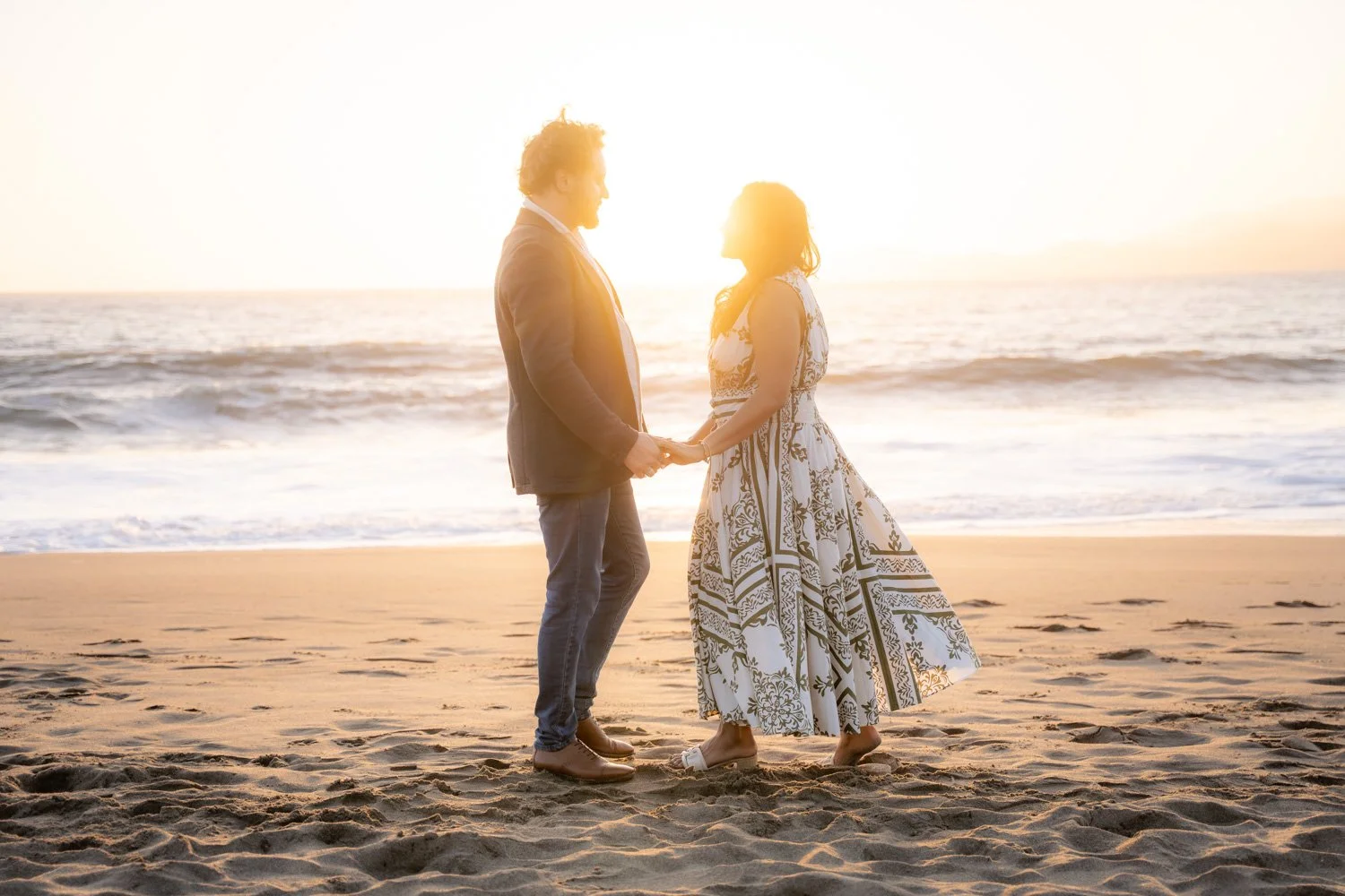 A couple holding hands on the beach at sunset, facing each other with the ocean in the background.