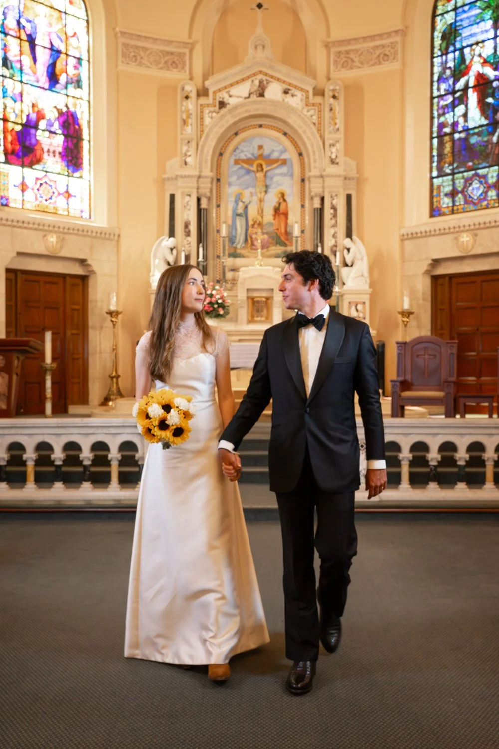 A bride and groom holding hands inside a church with stained glass windows and religious artwork in the background.