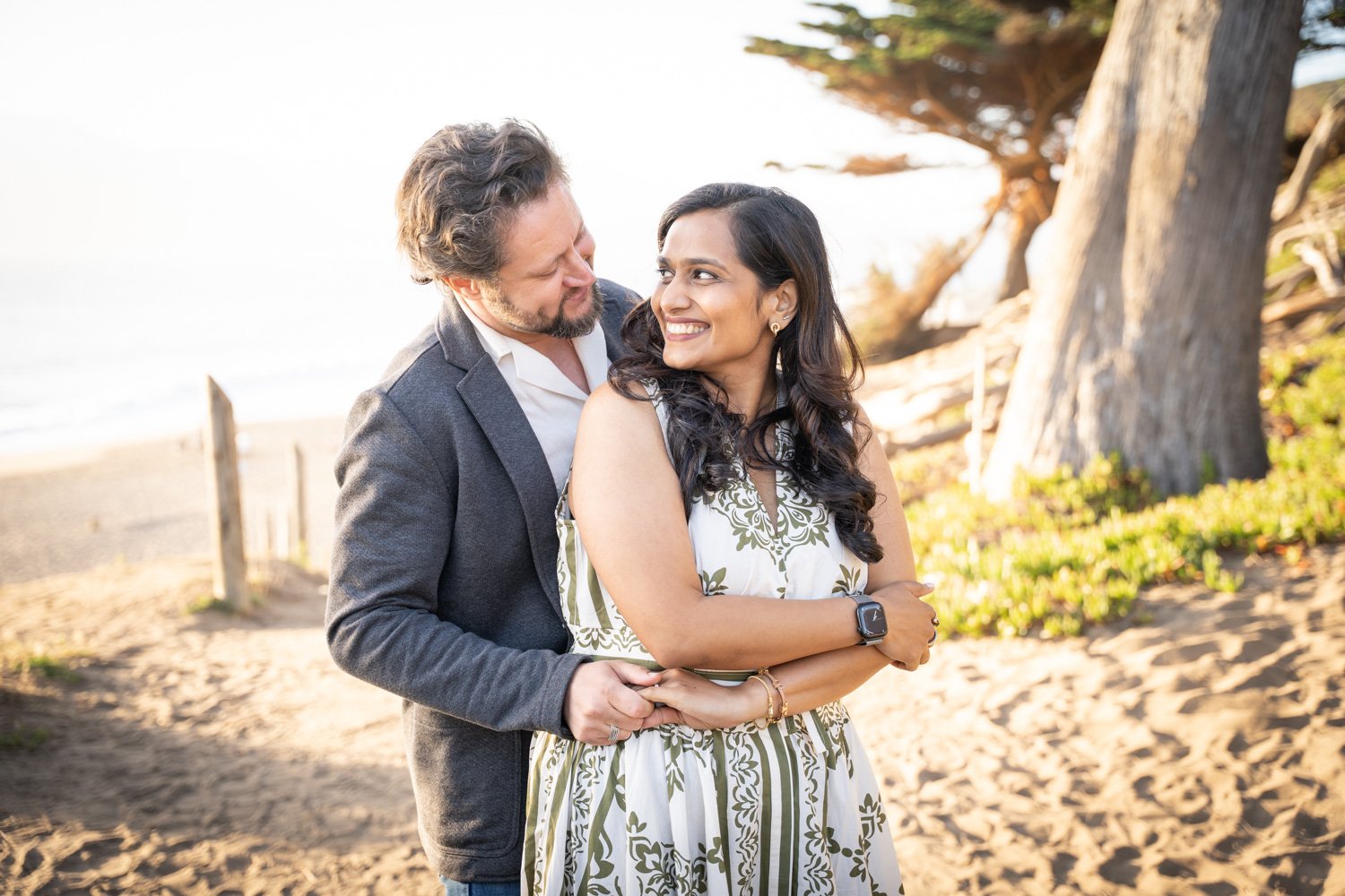 A couple standing on a sandy beach near trees, smiling at each other during sunset, with the man embracing the woman from behind.