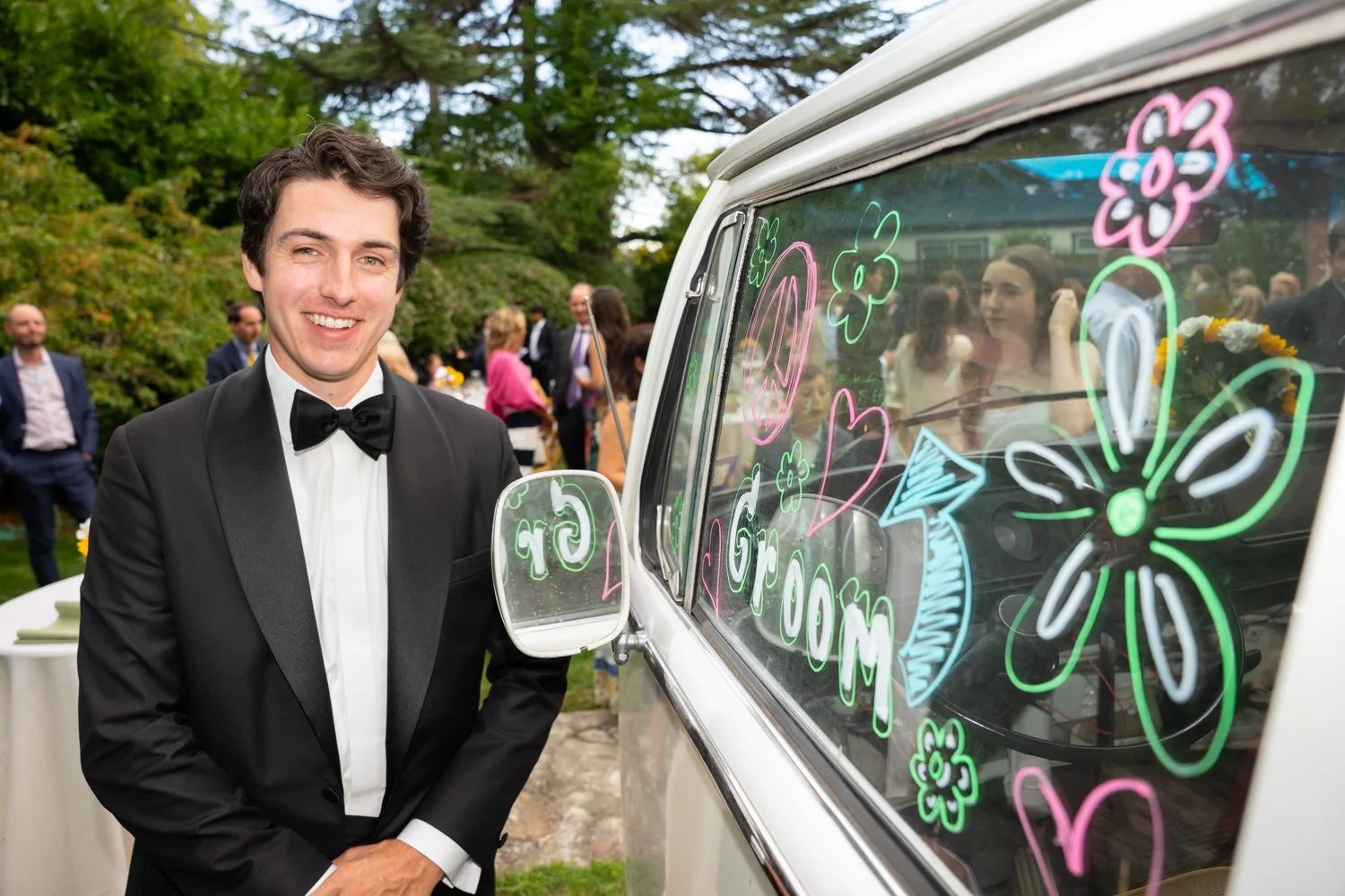 A man in a tuxedo stands next to a decorated vehicle with colorful flowers and hearts, at an outdoor event with other people in the background.