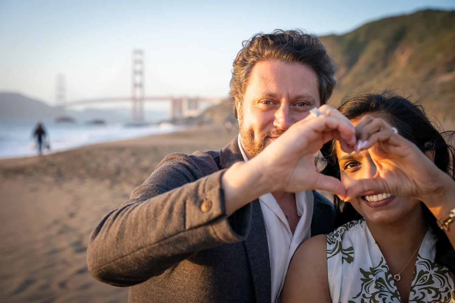 A happy couple forming a heart shape with their hands on a beach with the Golden Gate Bridge in the background.