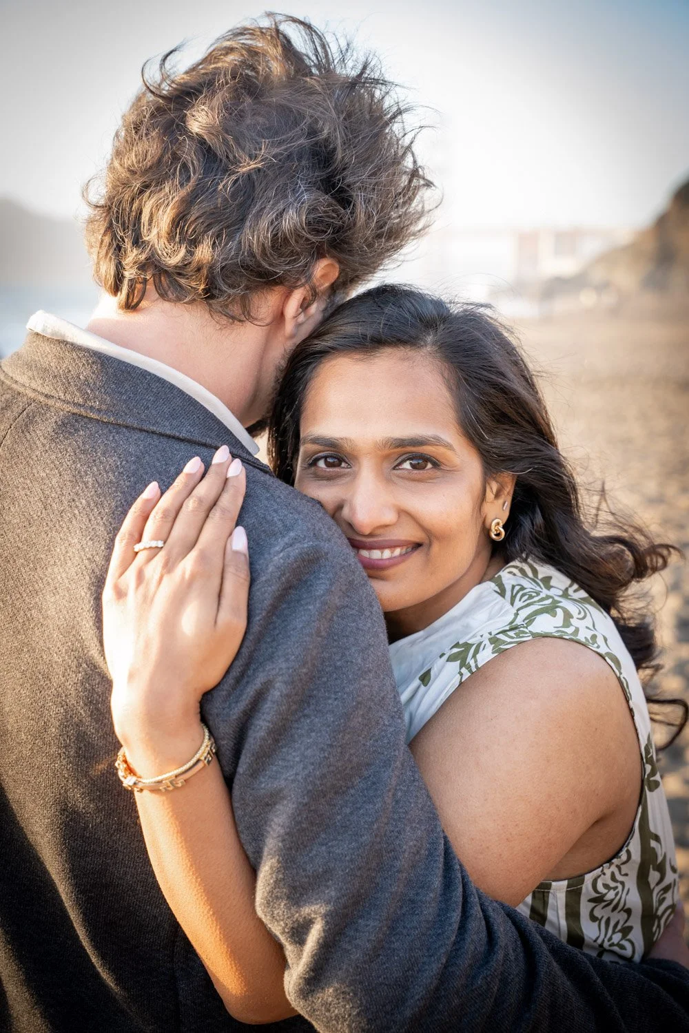 A romantic couple hugging on a beach at sunset, the woman smiling and looking into the camera, the man with his back to the camera.