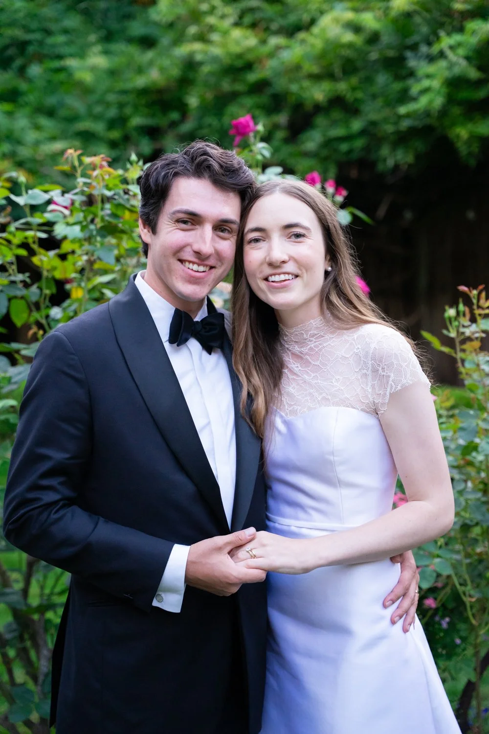 A young couple dressed in formal wedding attire, standing outdoors in front of green bushes and pink flowers, smiling at the camera.