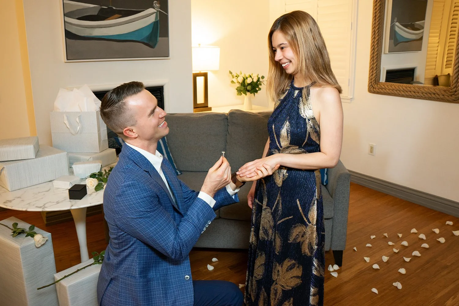 A man proposing marriage to a woman in a living room decorated for a romantic occasion with rose petals and gifts. The man is kneeling, holding a ring, and smiling at the woman. The woman is standing, smiling, and holding the man's hand.