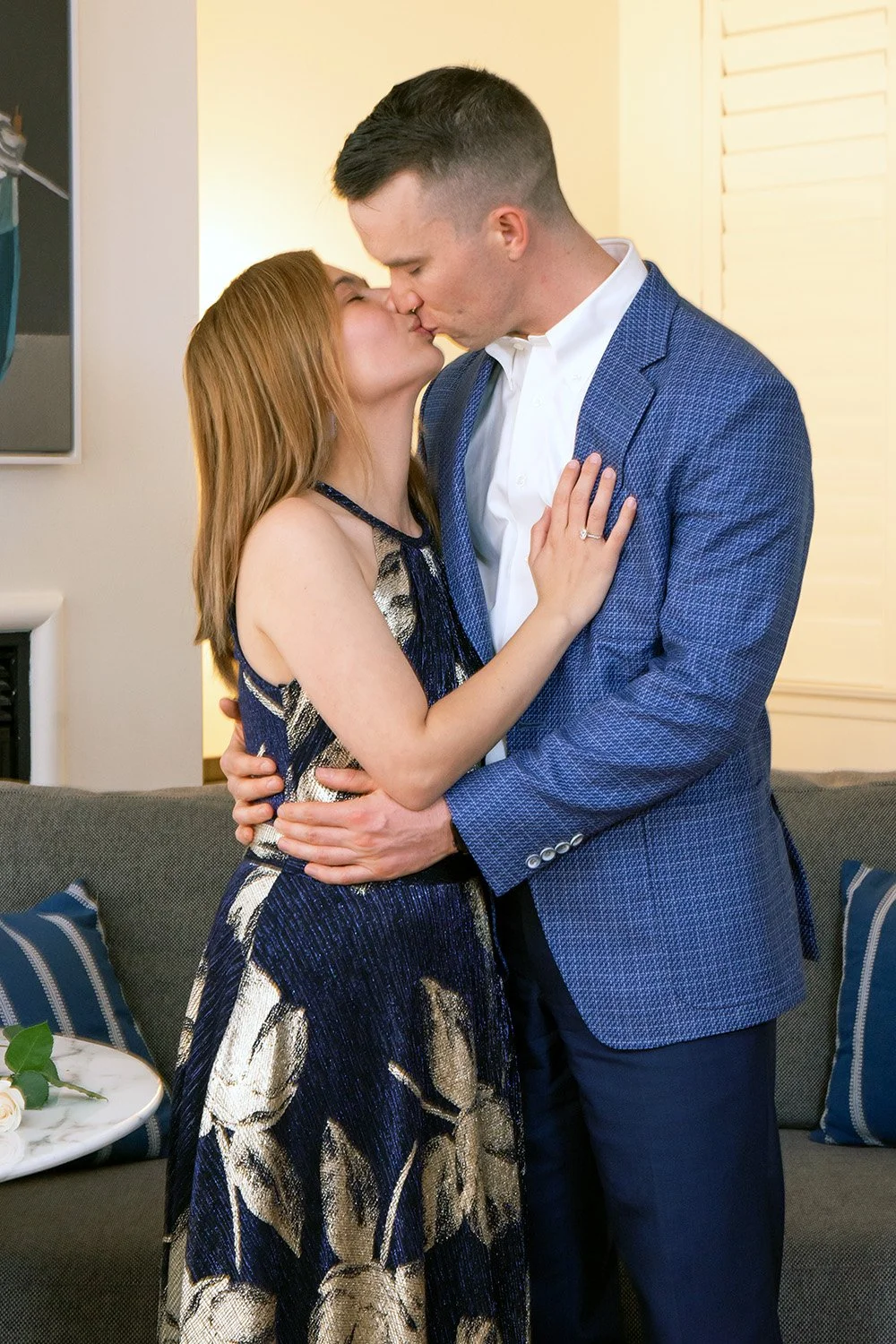 A couple sharing a kiss indoors, with a woman wearing a patterned dress and a man in a blue suit jacket and white shirt.
