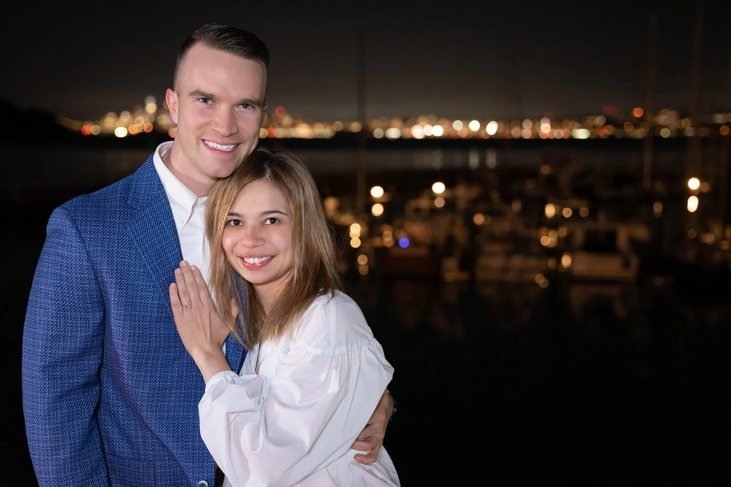 A smiling young couple, the man in a blue blazer and white shirt, the woman in a white blouse, embracing at night with city lights and boats in the background.