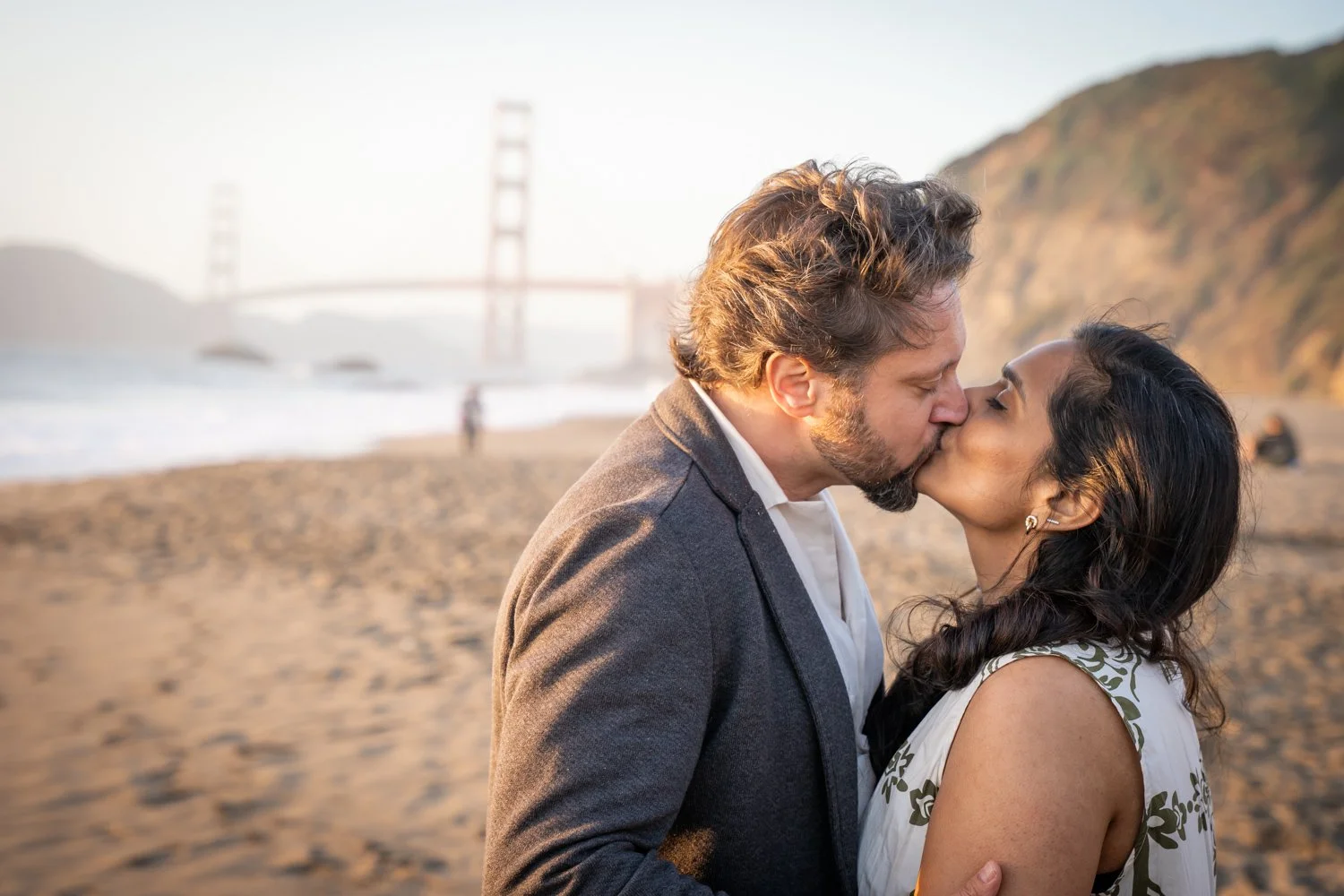 A couple kissing on a beach near the ocean with the Golden Gate Bridge in the background.