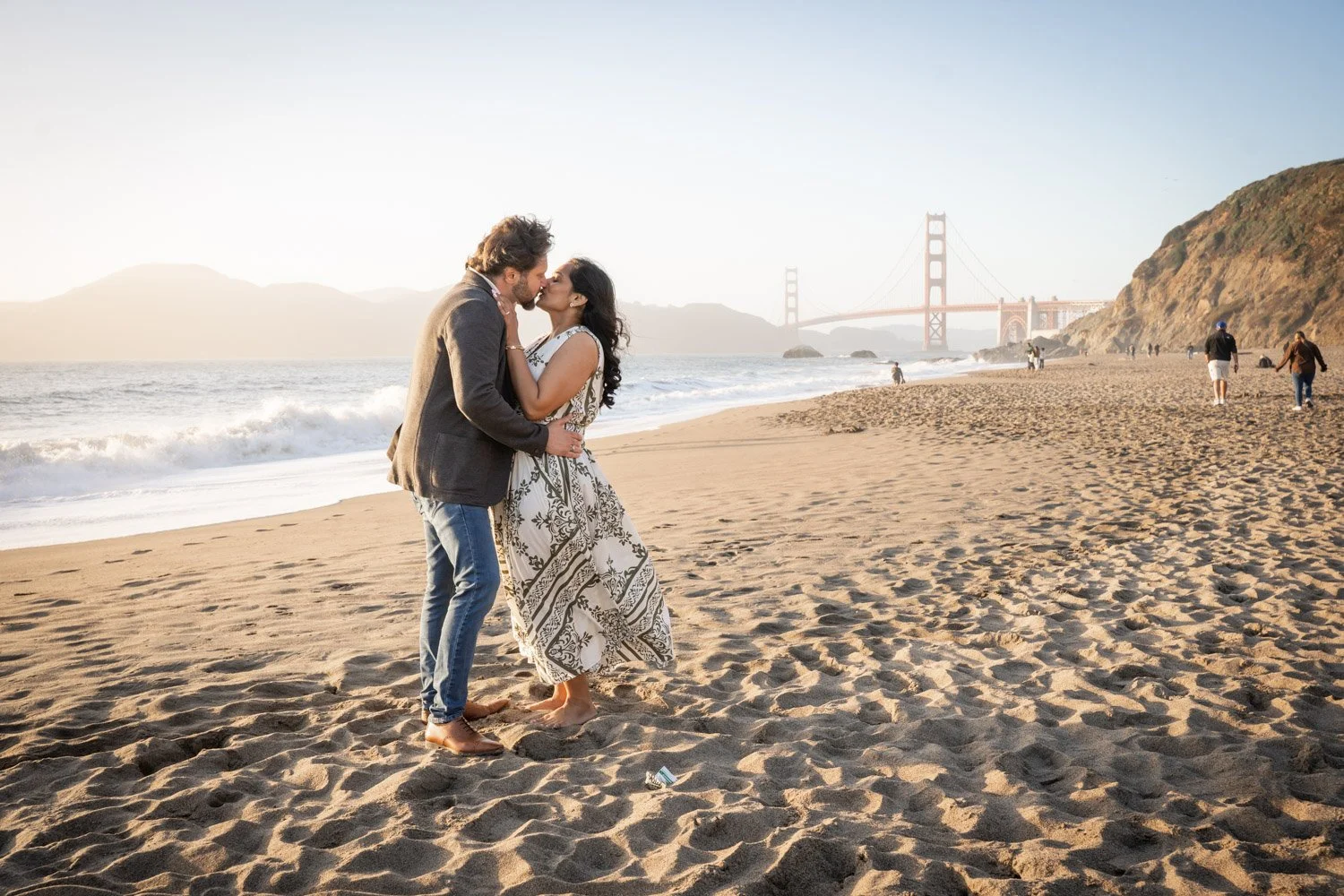 A man and woman kissing on a sandy beach with the Golden Gate Bridge in the background during sunset.
