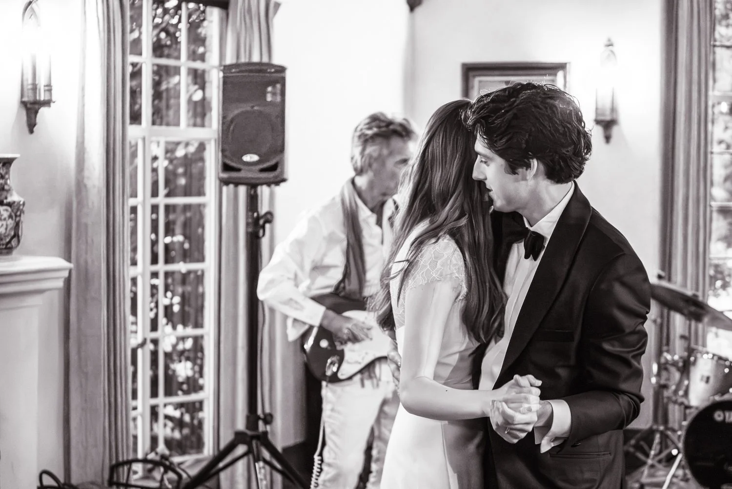 A young couple dancing closely at a wedding reception, with a musician playing guitar in the background, in a decorated indoor venue.