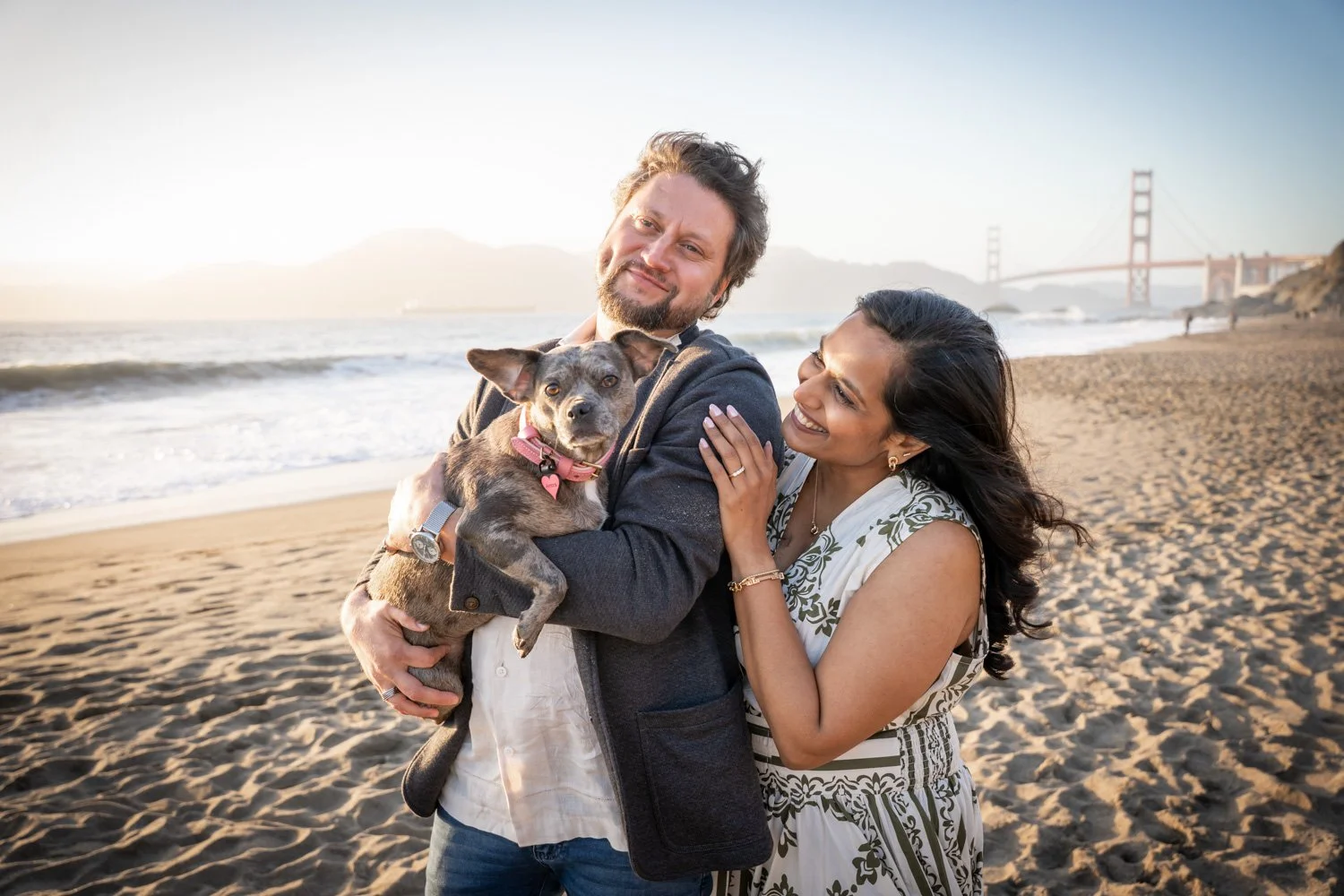 A happy couple and their dog on the beach near the Golden Gate Bridge at sunset.
