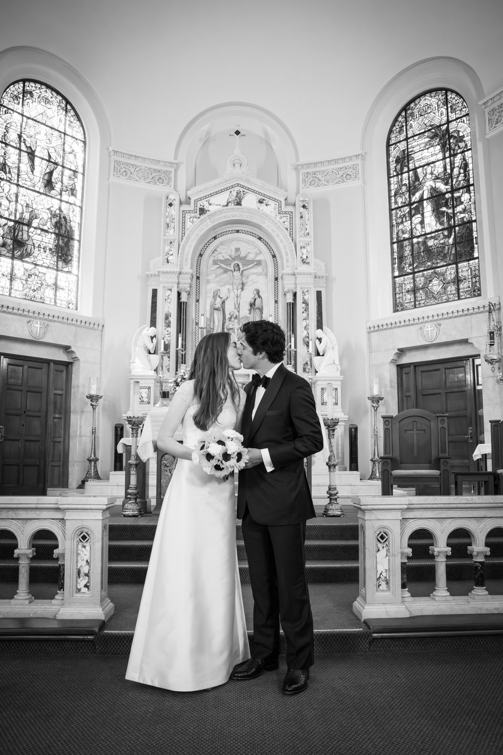 A bride and groom share a kiss inside a church, with the bride holding a bouquet of flowers and the groom in a tuxedo. The church features stained glass windows and religious statues.
