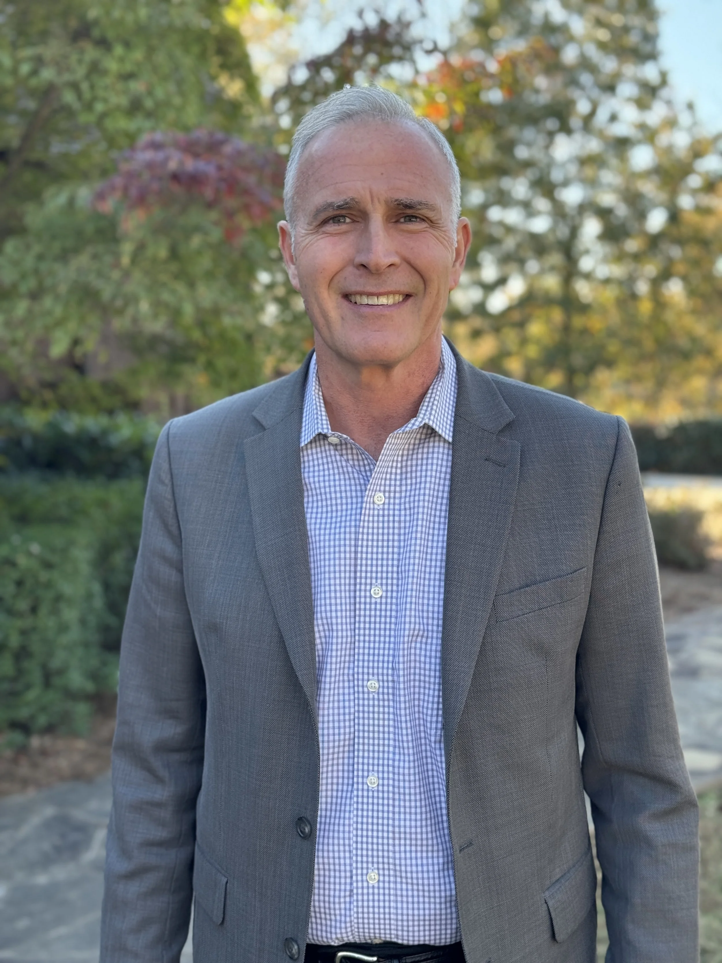 A middle-aged man with short gray hair, dressed in a gray suit jacket and a checkered dress shirt, standing outdoors in a park with trees and foliage in the background.