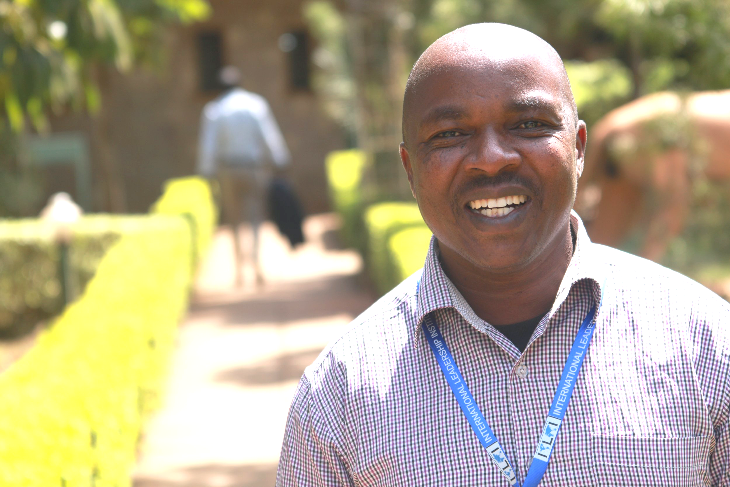Smiling man with a checkered shirt and an international leadership badge in an outdoor setting with greenery and a blurred background.