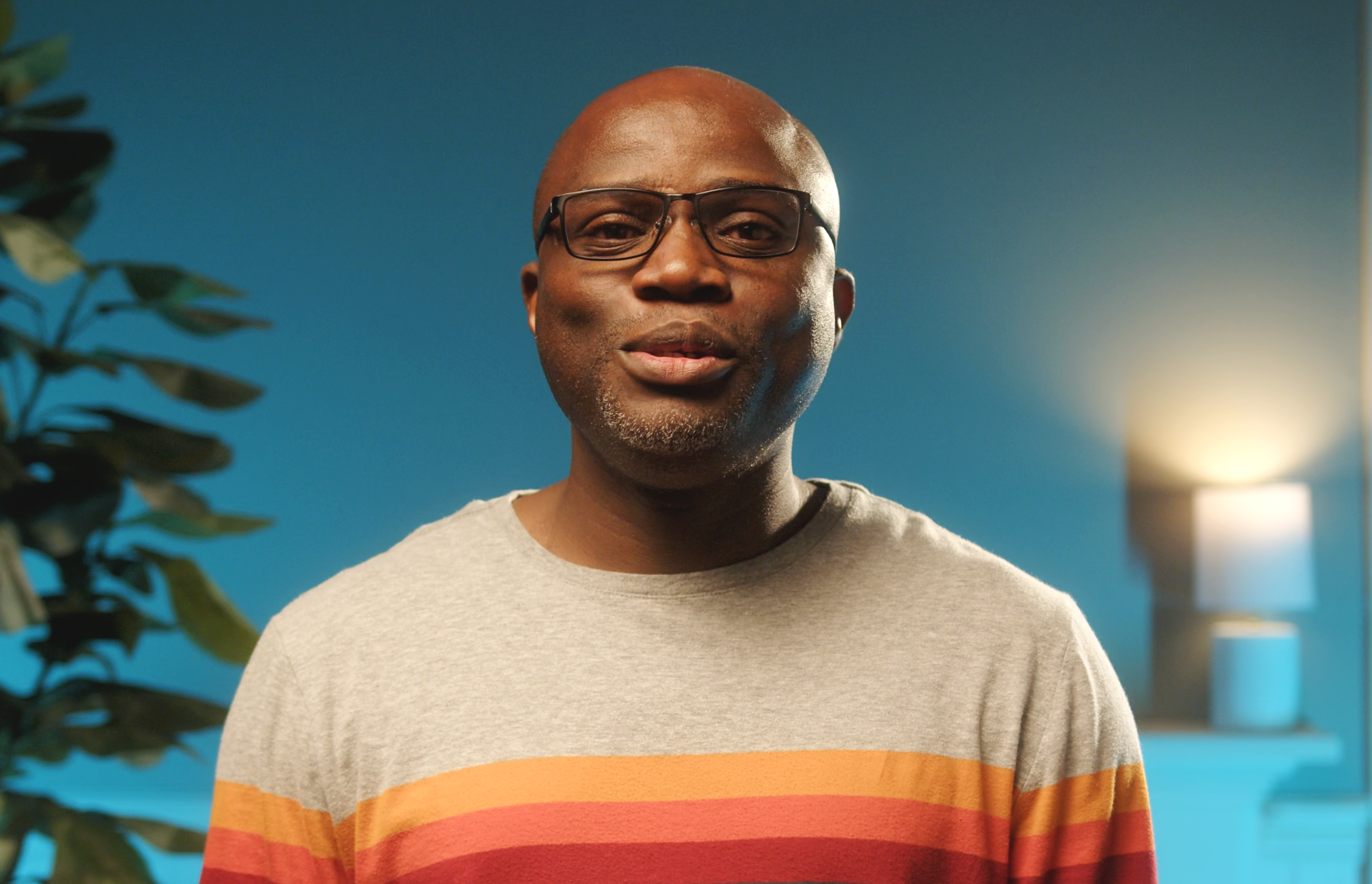 Man wearing glasses and a striped shirt, standing indoors with a blue wall and a lamp in the background.