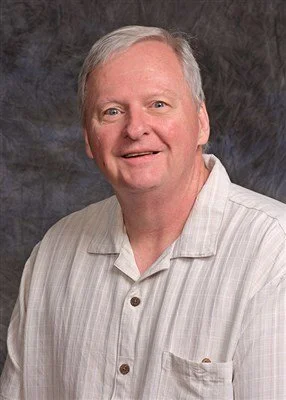 A smiling man with gray hair wearing a light-colored, short-sleeved, collared shirt.