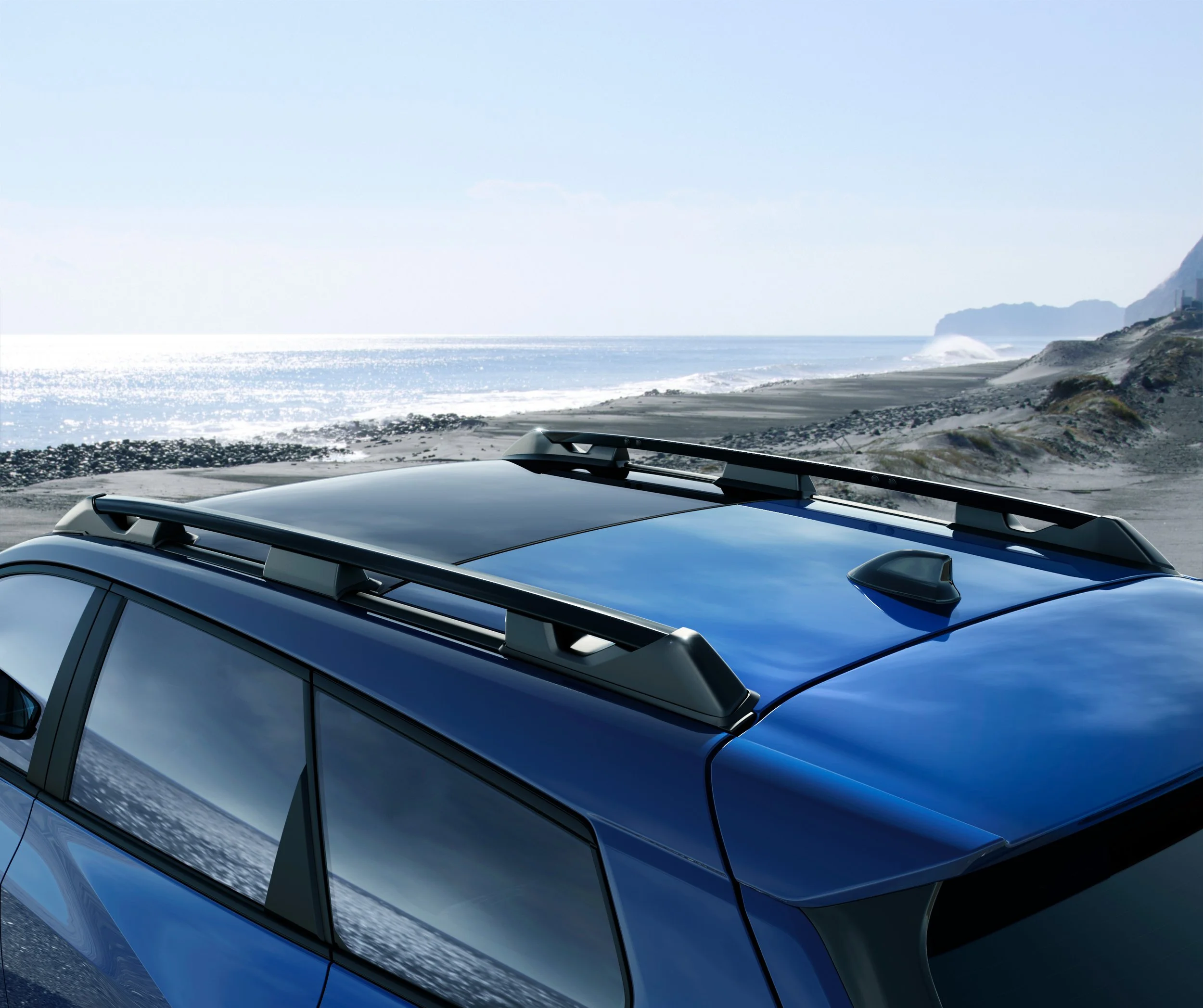 Nærbilde av taket på en blå bil med takbøyler ved en kystlinje med sand og stein, sjø og klipper i bakgrunnen.