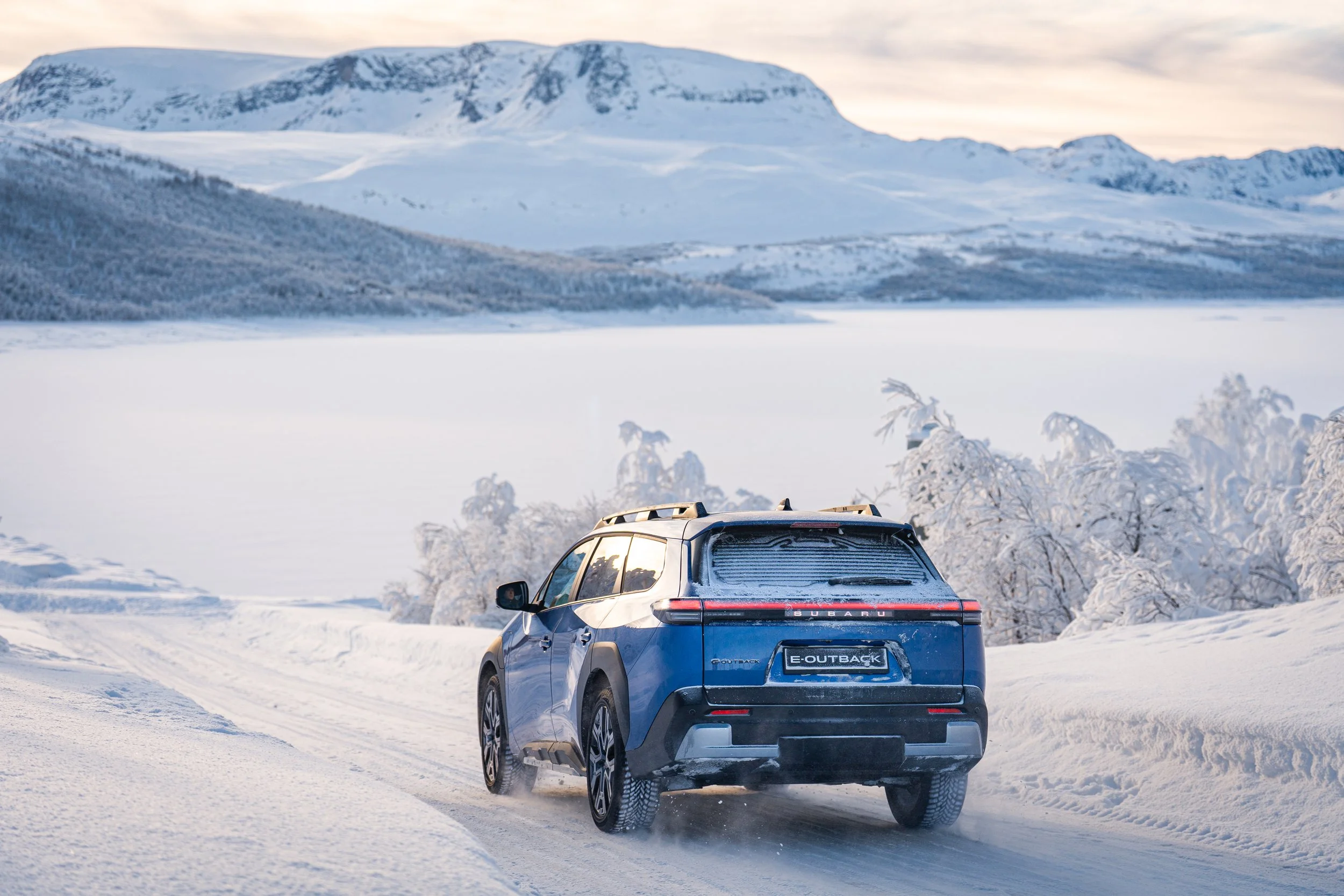 En blå Subaru Outback kjører på en snødekt vei i snødekte landskap med fjell i bakgrunnen. E-Outback fås hos Aaland bil, Ålesunds merkeforhandler og merkeverksted av Subaru.