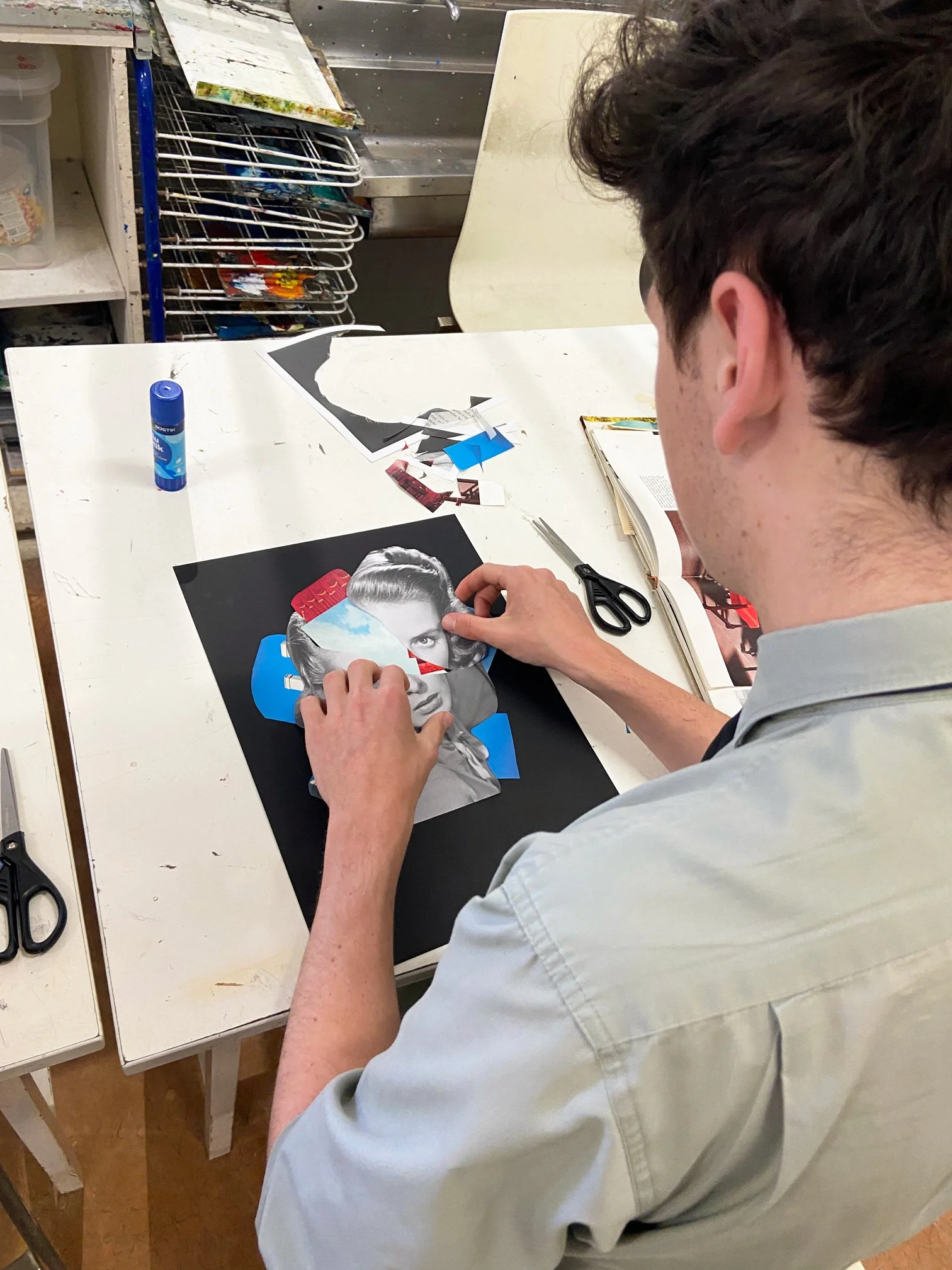 A Scots college student working on a collage at a white table, with scissors, glue stick, and paper scraps around, using black and white magazine photos, colored paper, and printed images.