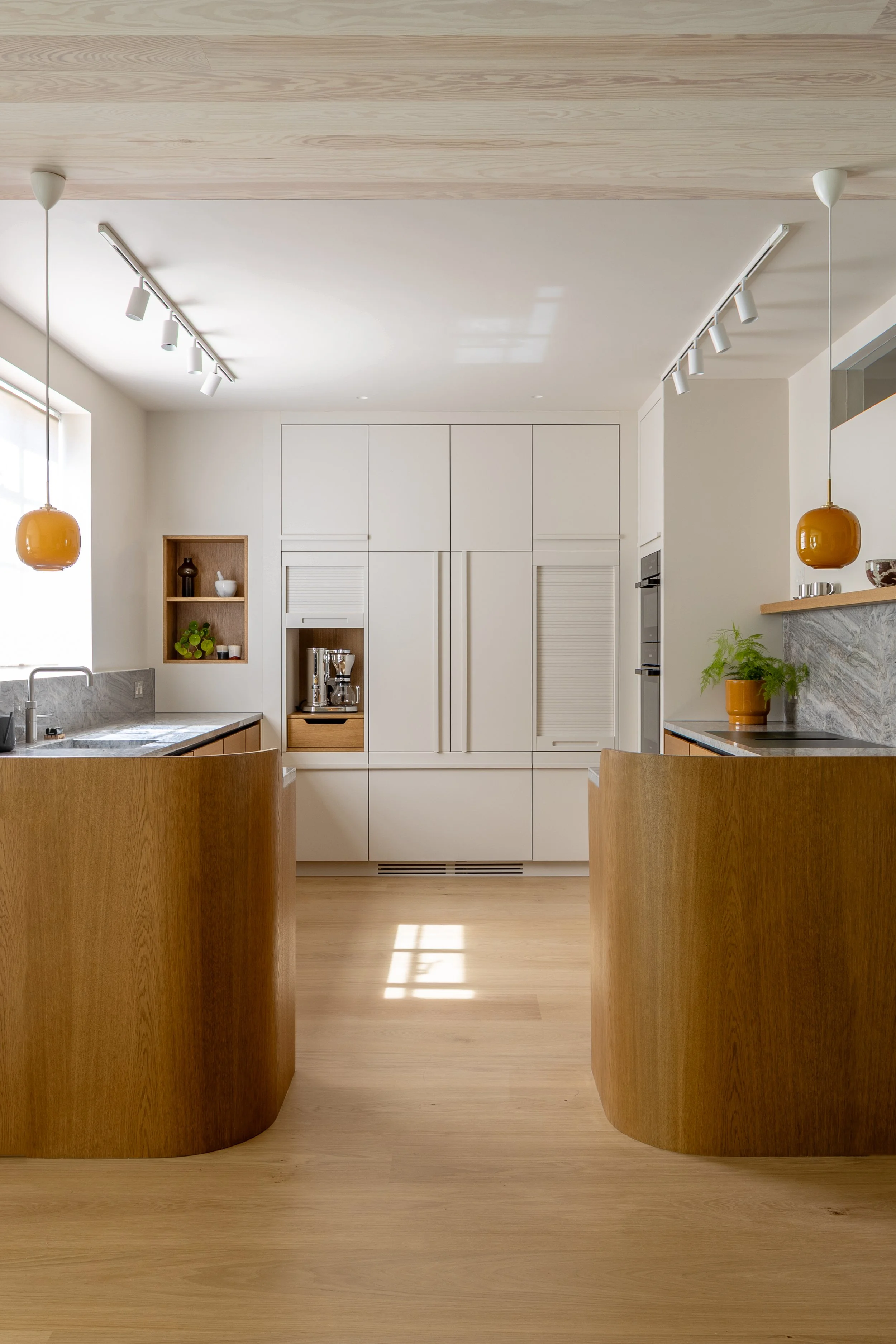 Bespoke kitchen in Islington by ALL & NXTHING, featuring curved oak cabinetry, grey marble worktops, full-height cream cupboards and amber pendant lights.