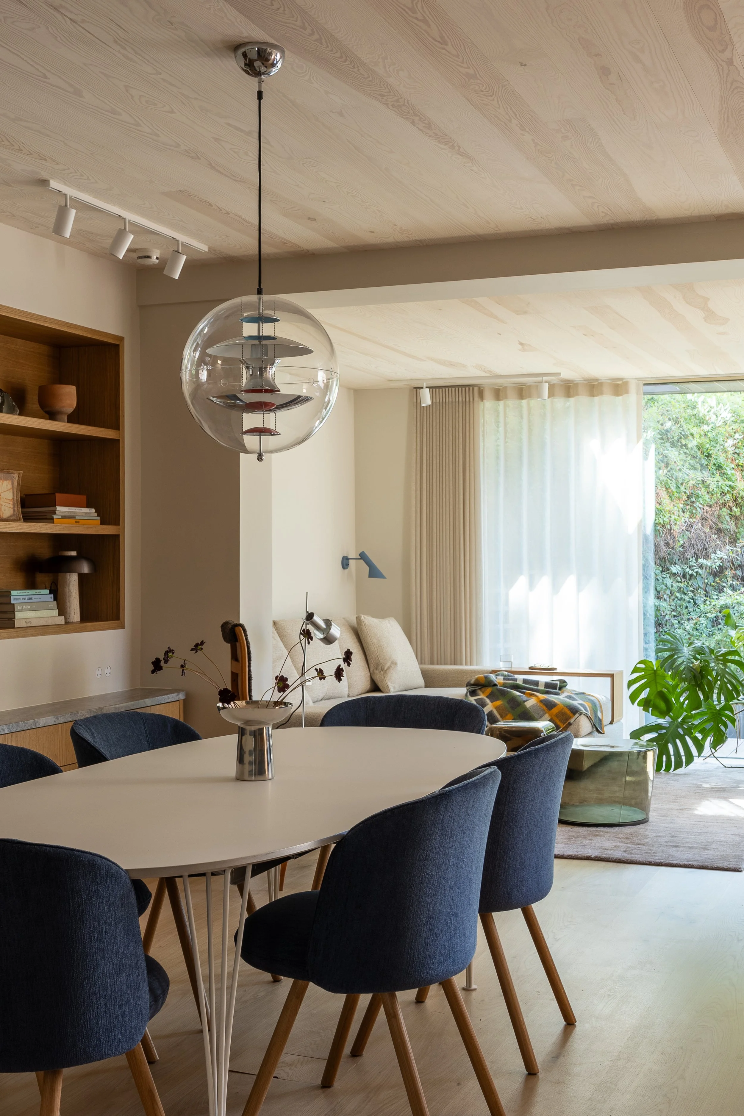 Open-plan dining and living room in Islington by ALL & NXTHING, with blue dining chairs, a glass globe pendant, timber ceiling and garden views.